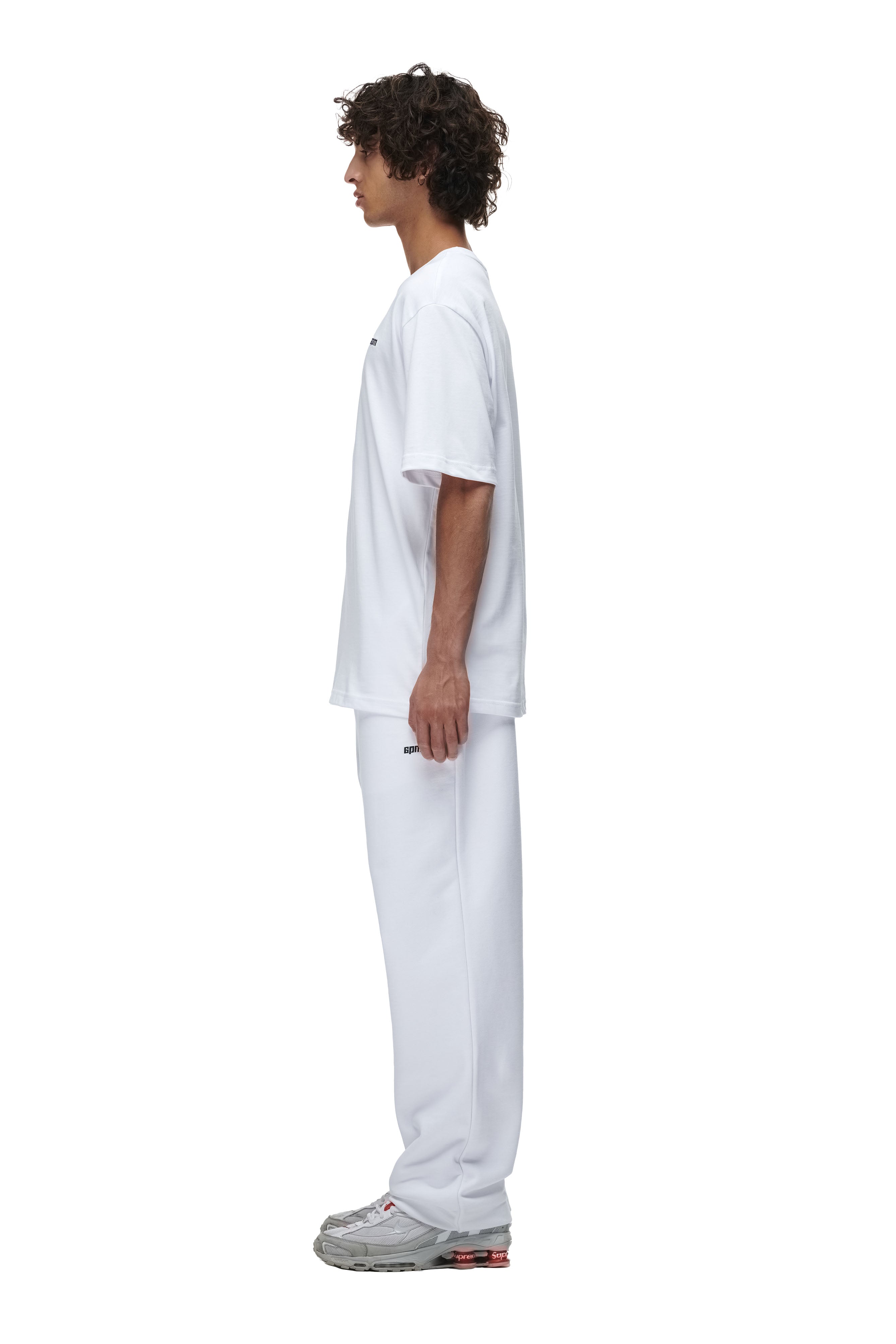 A person with curly hair stands in profile, facing left, wearing the 6PM T-SHIRT WHITE with matching white sweatpants and gray sneakers against a plain white background.