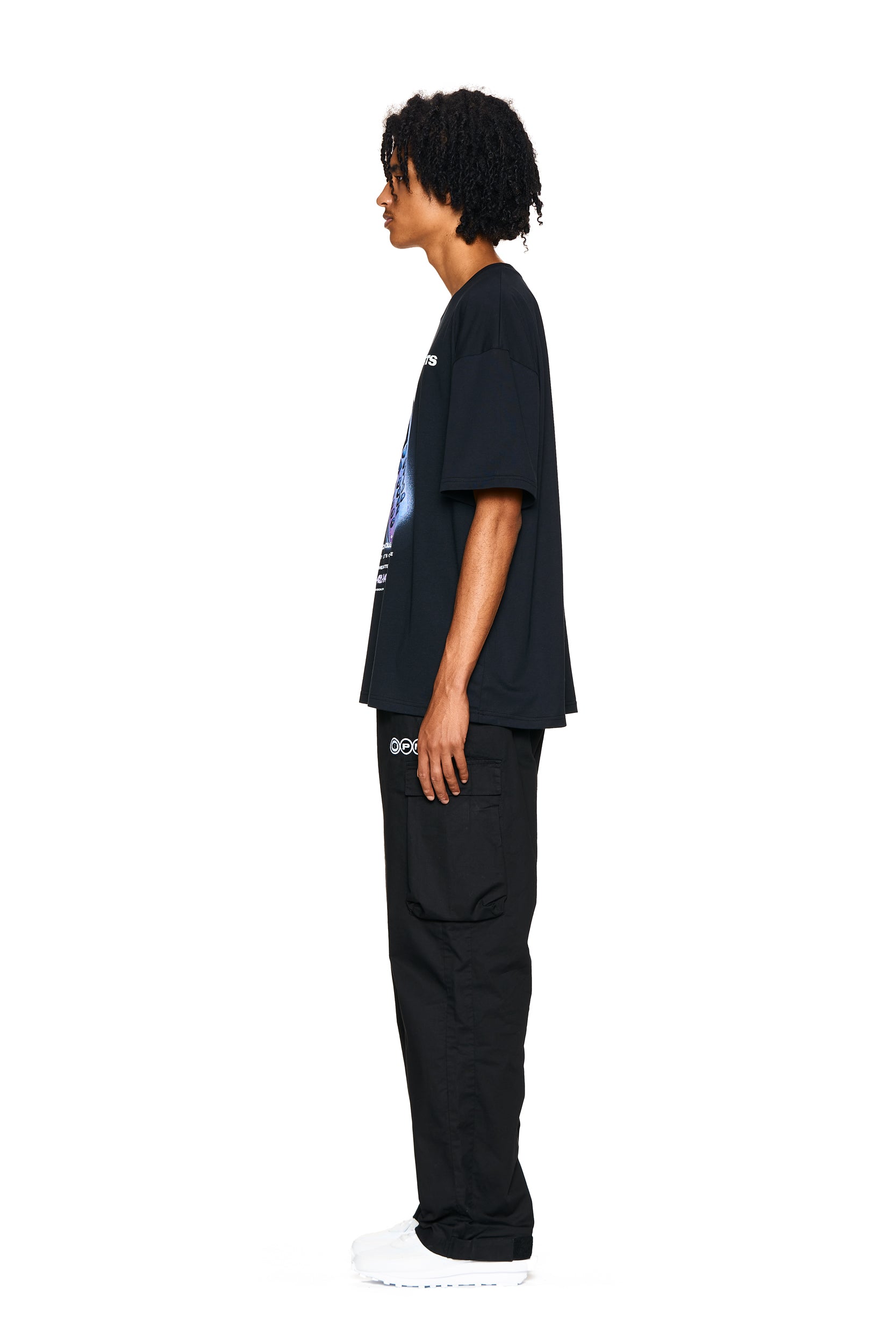 A man with curly hair stands in profile against a white background, wearing the 6PM PHONE T-SHIRT BLACK, black cargo pants with pockets, and white sneakers.