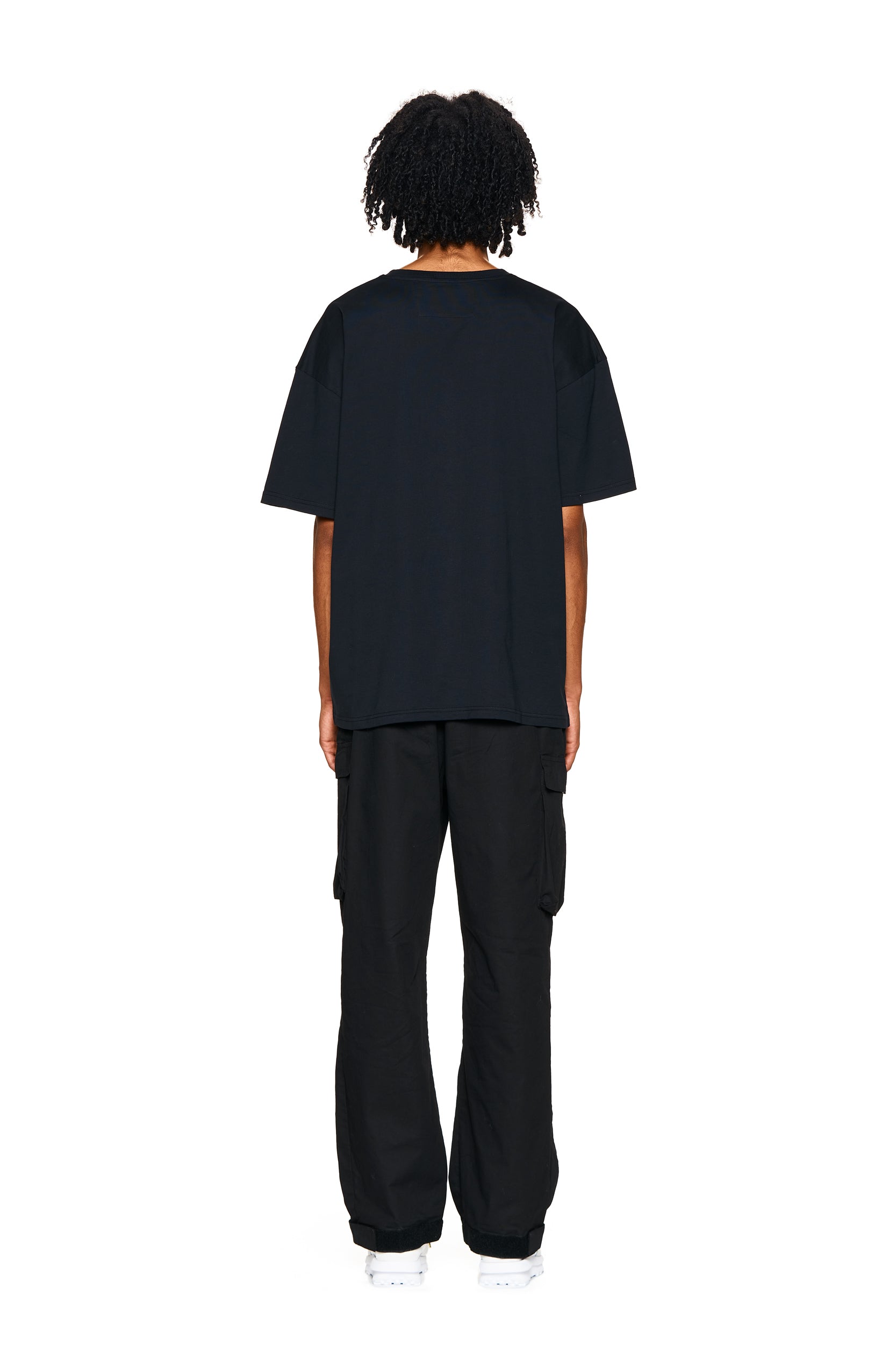 A person with curly hair stands facing away, wearing the 6PM PHONE T-SHIRT BLACK, black cargo pants, and white sneakers against a plain white background.