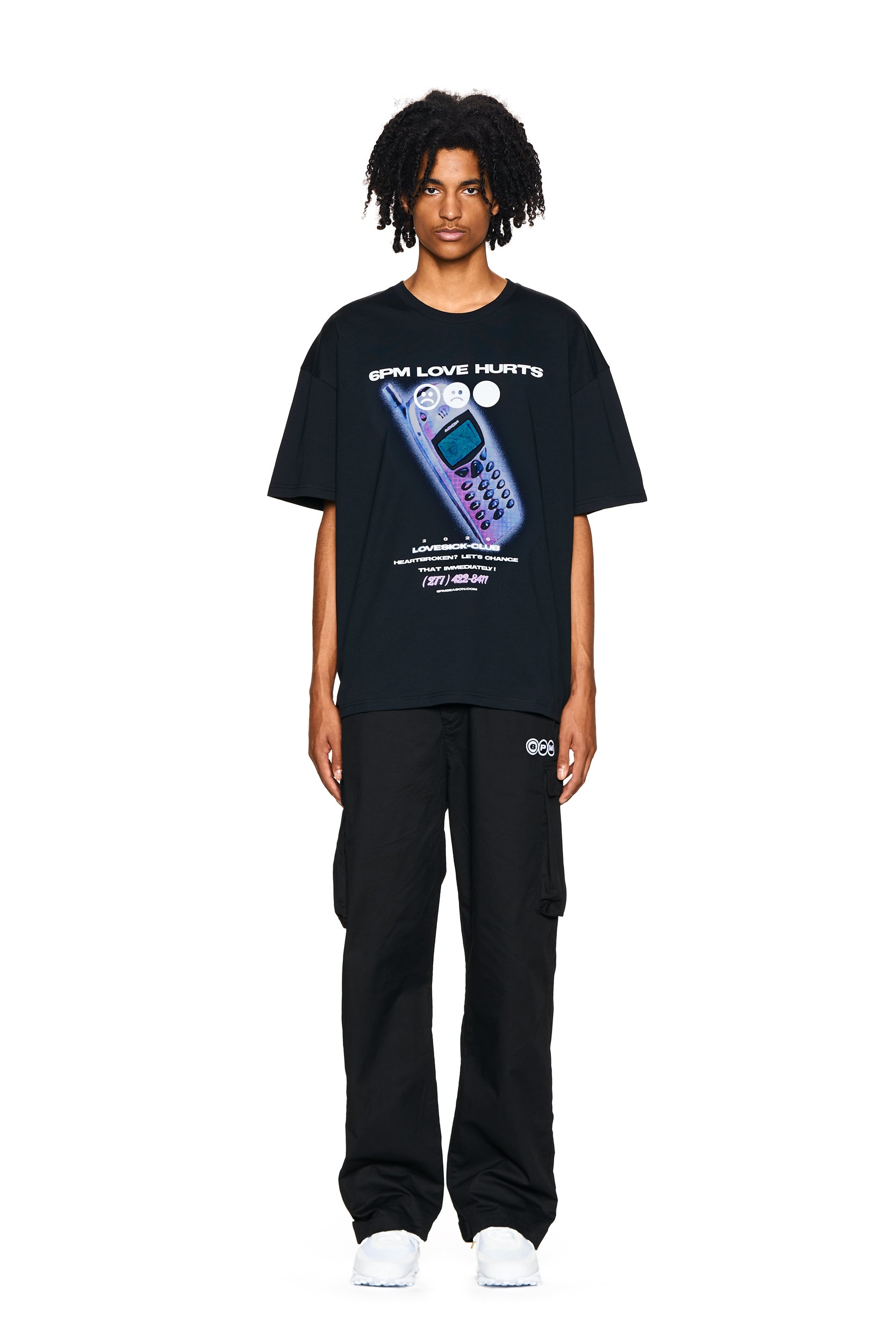 A person with medium-length curly hair wears the 6PM PHONE T-SHIRT BLACK featuring a retro cellphone graphic, paired with black cargo pants and white sneakers, standing against a plain white background.