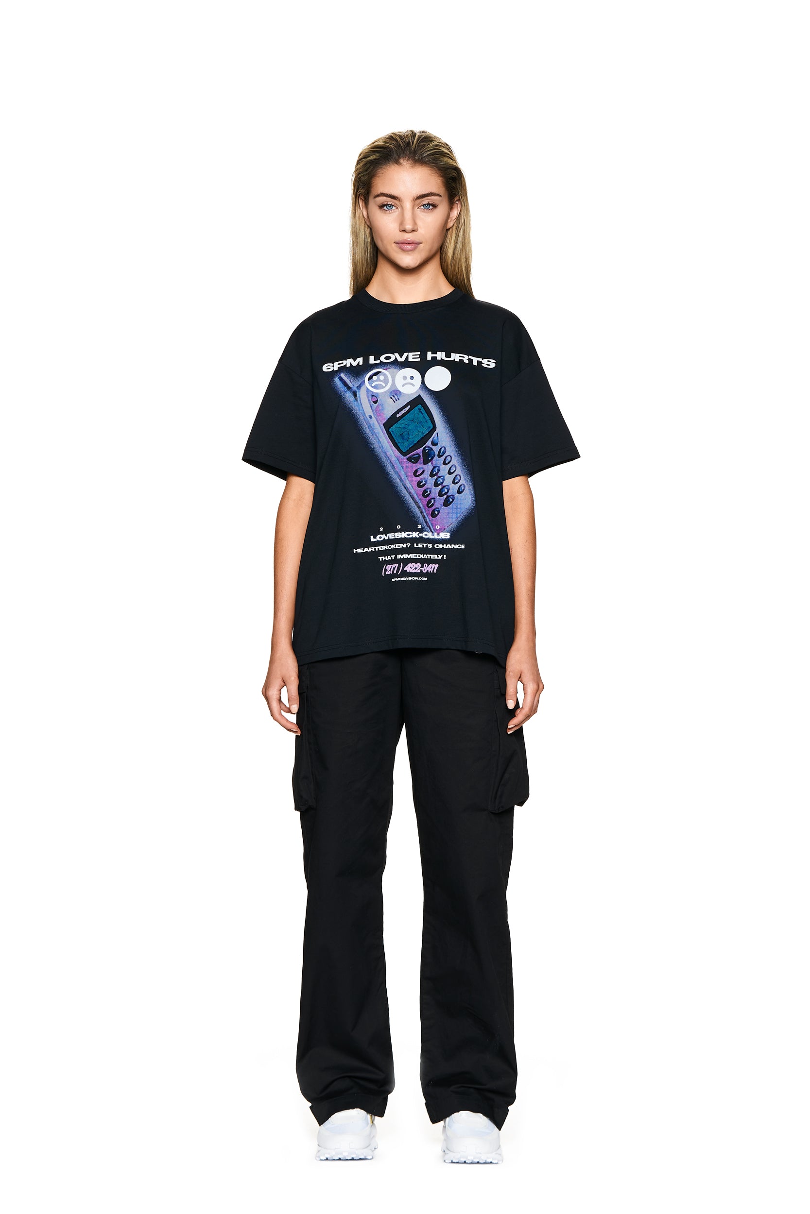 A woman faces forward in a plain white setting, wearing the 6PM PHONE T-SHIRT BLACK featuring "LOVE HURTS" with a cell phone graphic, paired with black cargo pants and white sneakers.