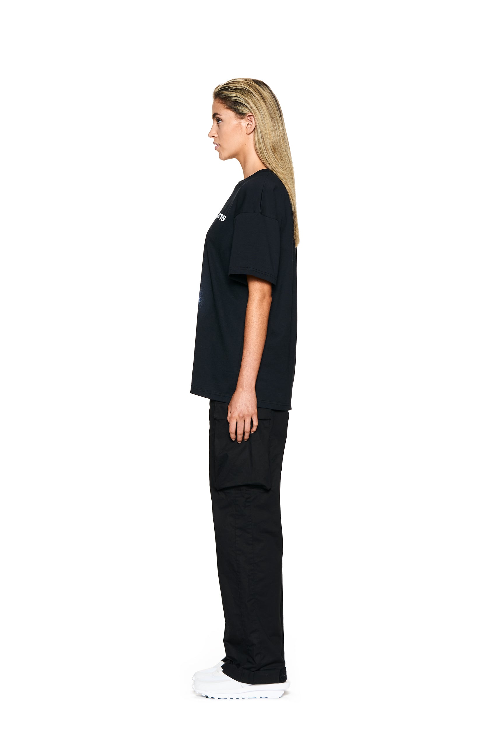 A woman with straight blonde hair stands in profile, facing left. She wears the 6PM PHONE T-SHIRT BLACK with black cargo pants and white sneakers, posed against a plain white background.