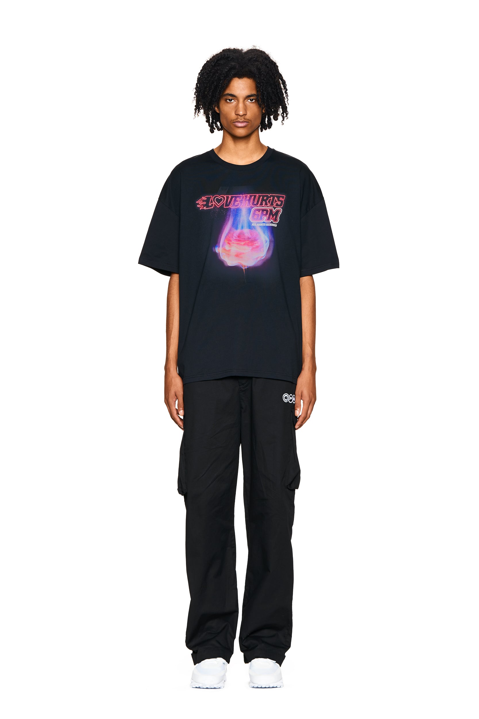 A person stands facing forward wearing the 6PM LOVE HURTS T-SHIRT BLACK, paired with black cargo pants and white shoes, against a plain white background.