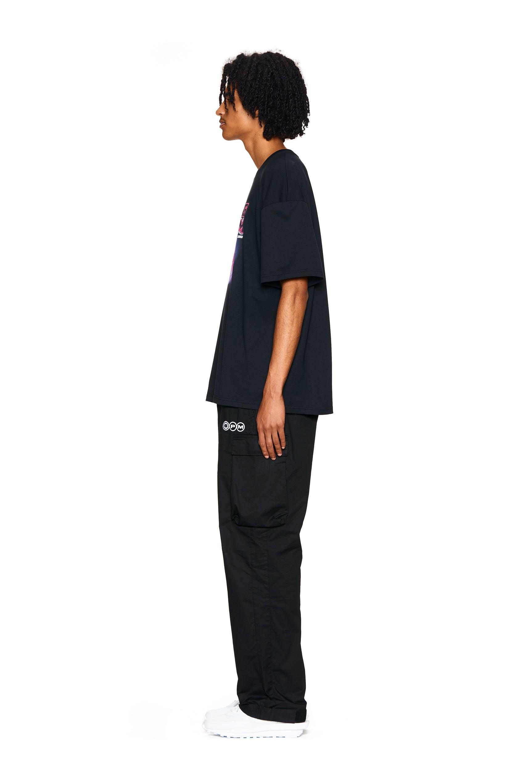A person with curly hair stands in profile against a white background, wearing the 6PM LOVE HURTS T-SHIRT BLACK, black cargo pants with a white logo on the pocket, and white sneakers.