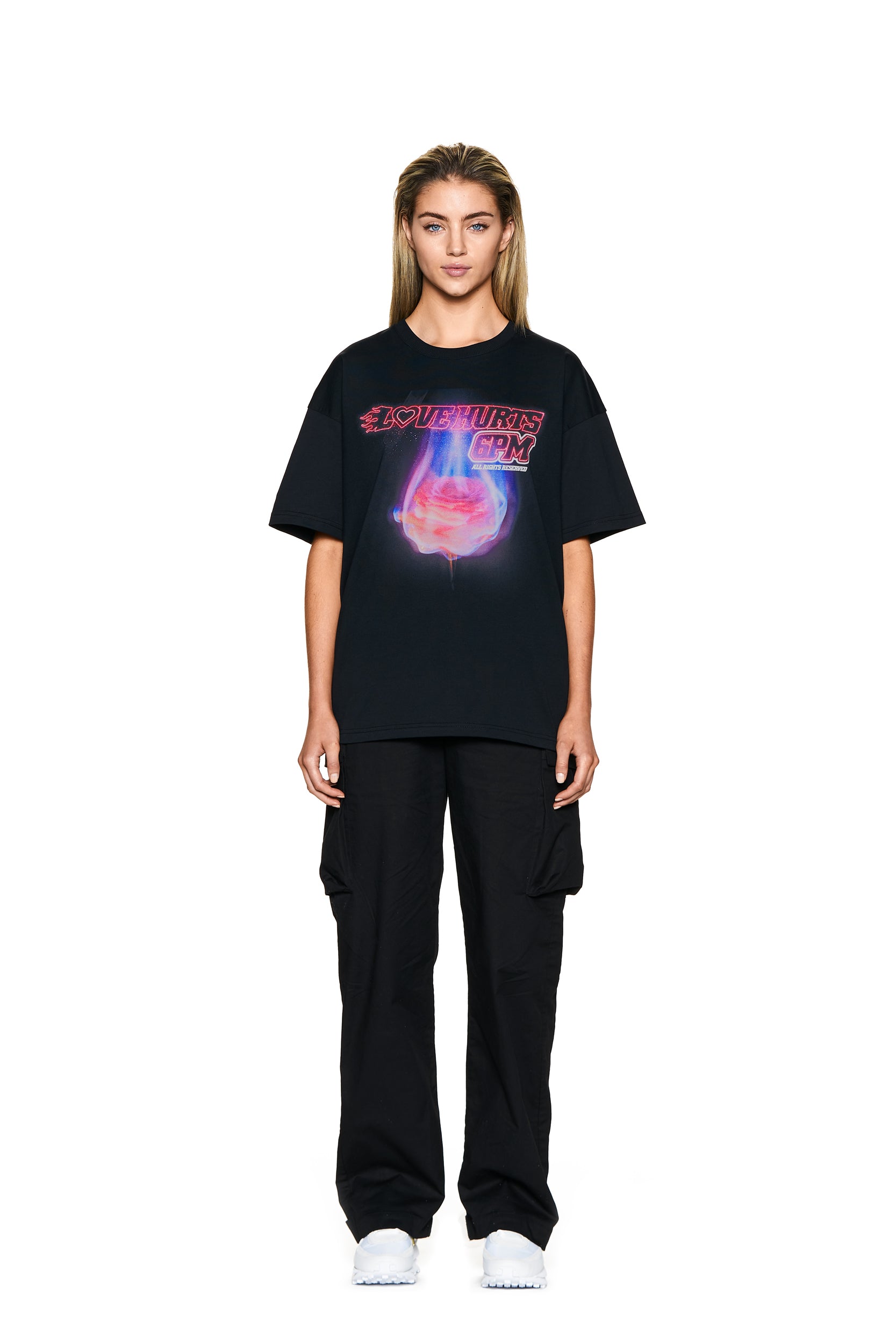 A woman with straight, light brown hair wears the 6PM LOVE HURTS T-SHIRT BLACK featuring purple-pink graphics, paired with black cargo pants and white shoes, standing on a plain white background.