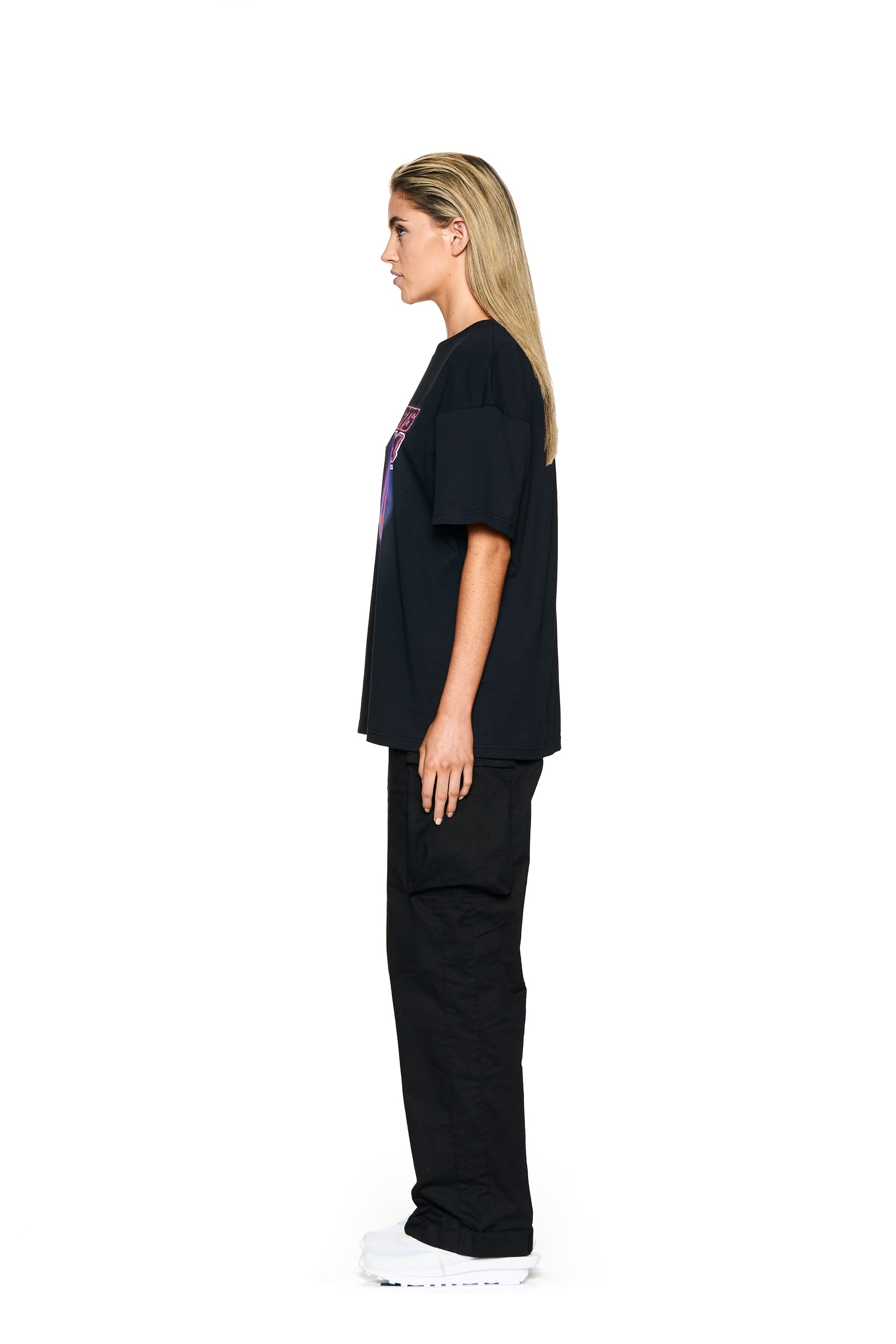 A woman with straight blonde hair stands in profile against a white background, wearing the 6PM LOVE HURTS T-SHIRT BLACK, black pants, and white sneakers.