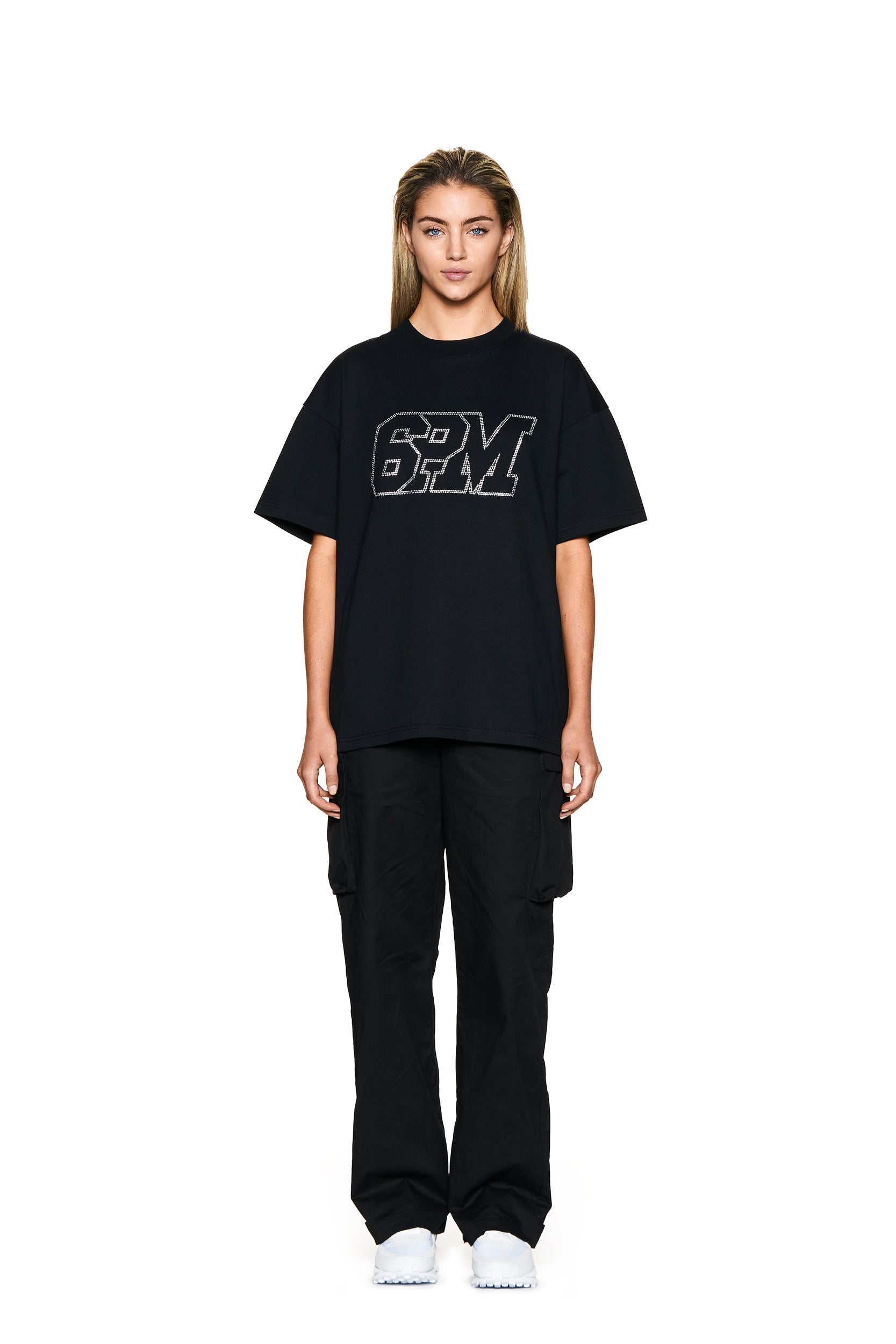 A woman with straight light brown hair faces forward against a white background, wearing the 6PM BLING T-SHIRT BLACK, black cargo pants, and white sneakers. She has a neutral expression.