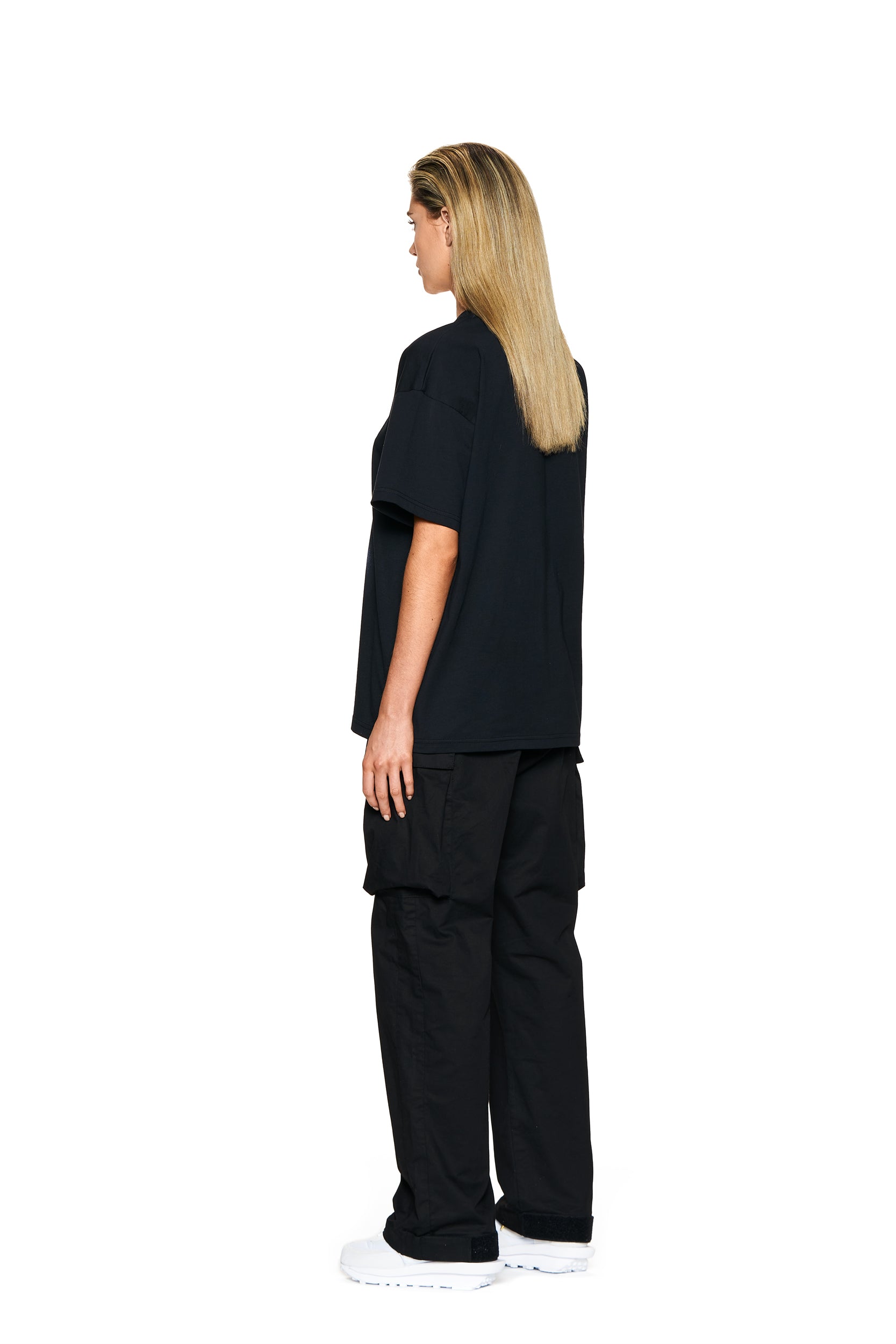 A person with straight, shoulder-length blonde hair stands sideways, facing left, wearing the 6PM BLING T-SHIRT BLACK, black cargo pants, and white shoes against a plain white background.