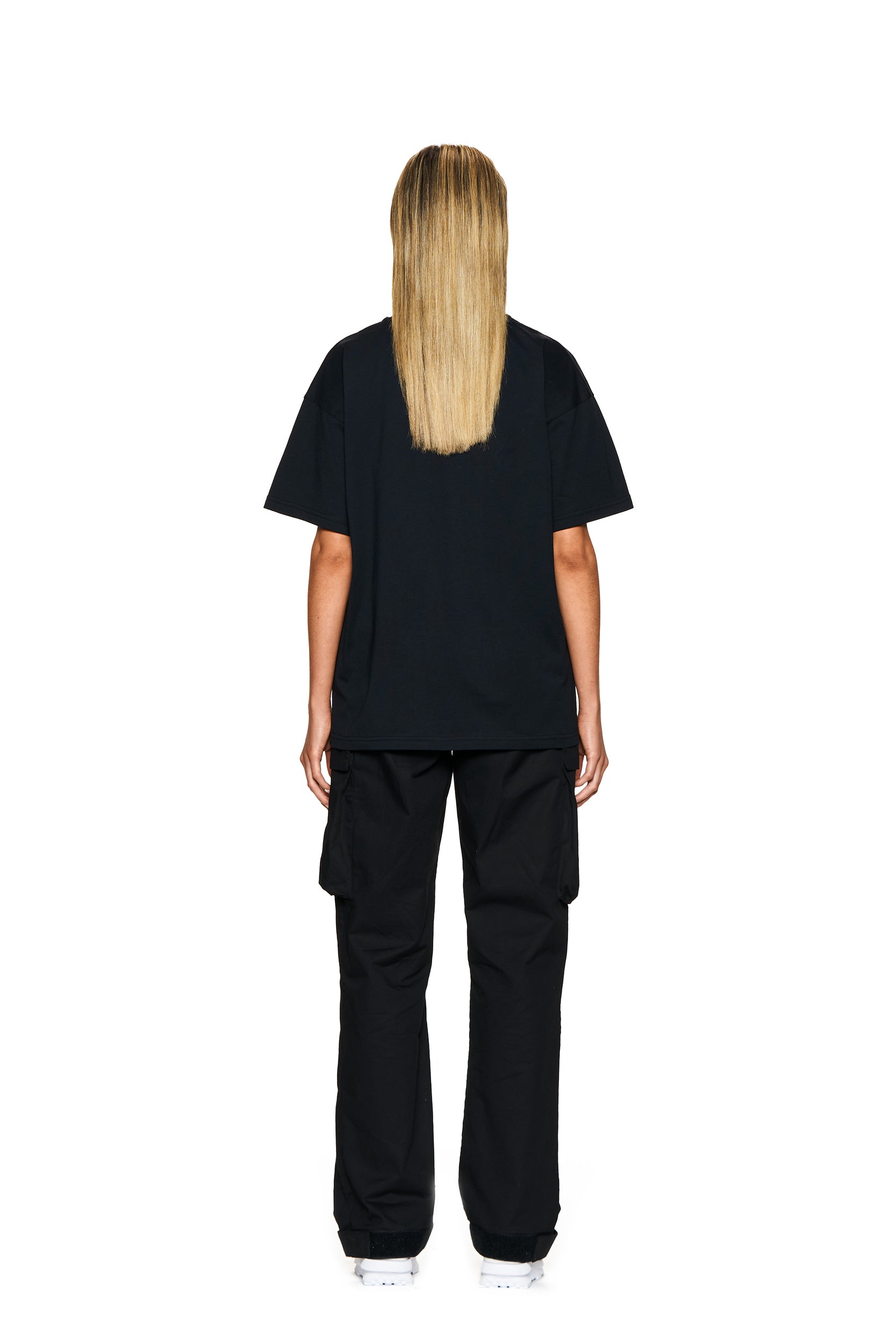 A person with long, straight blonde hair faces away from the camera, wearing the 6PM BLING T-SHIRT BLACK, black cargo pants, and white sneakers against a plain white background.