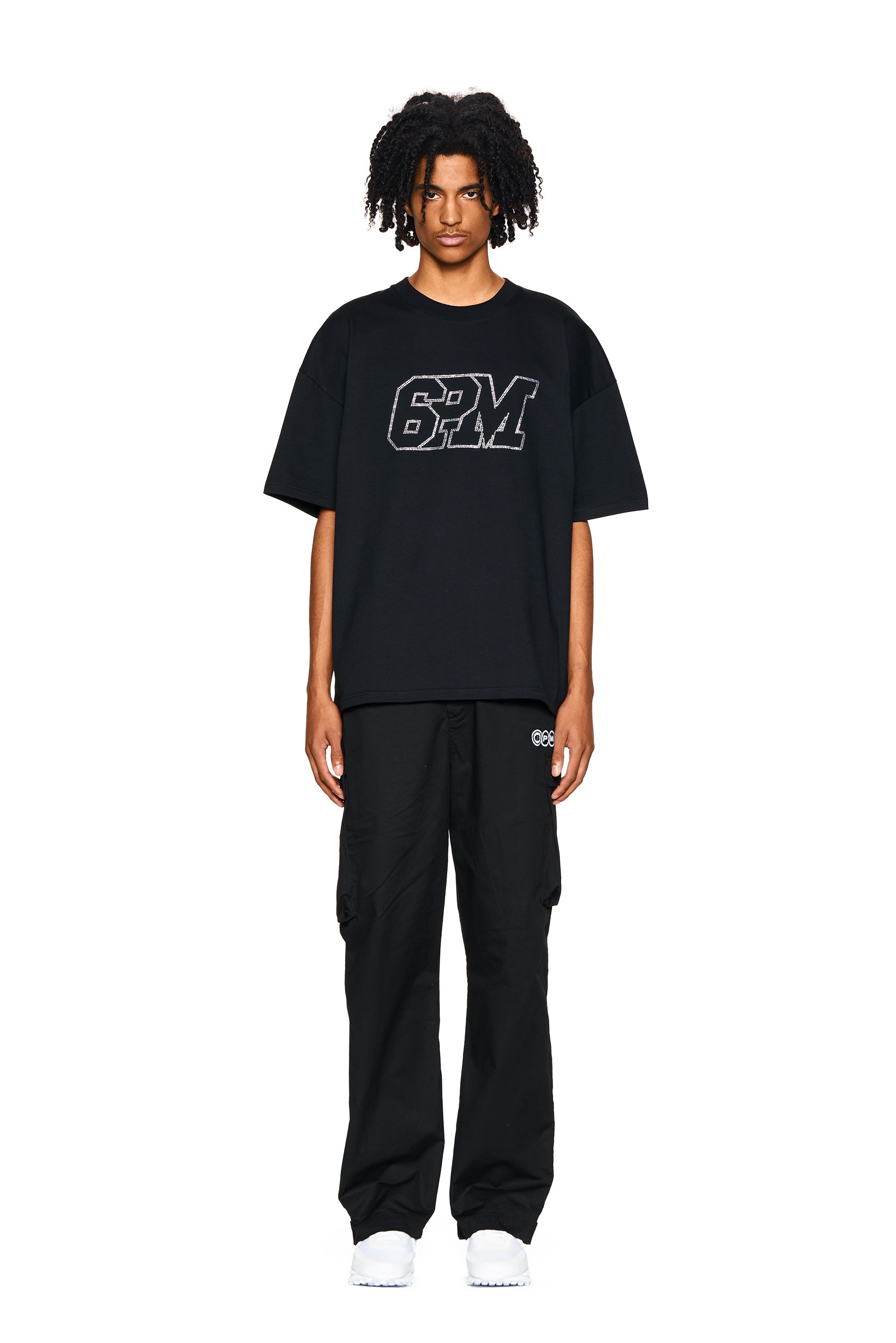 A person with medium-length curly hair faces forward, wearing the 6PM BLING T-SHIRT BLACK, black cargo pants, and white sneakers against a plain white background.