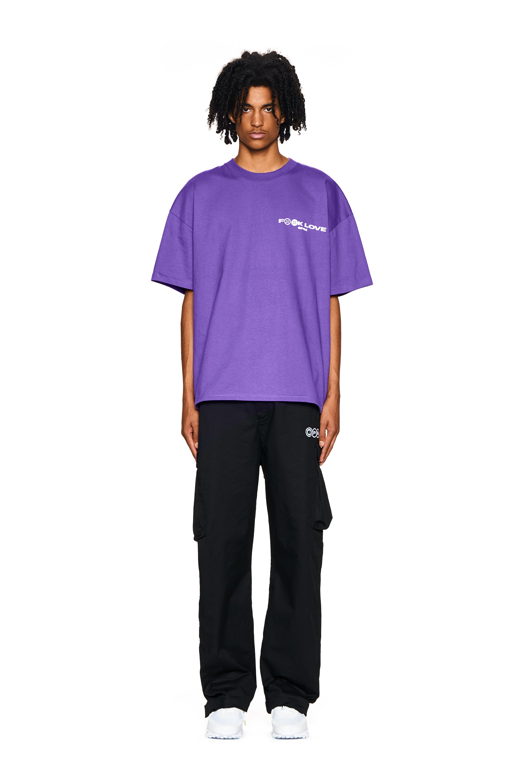 A person with medium-length curly hair and a neutral expression stands facing forward against a plain white background, wearing the 6PM FUCK LOVE T-SHIRT PURPLE, black cargo pants, and white sneakers.