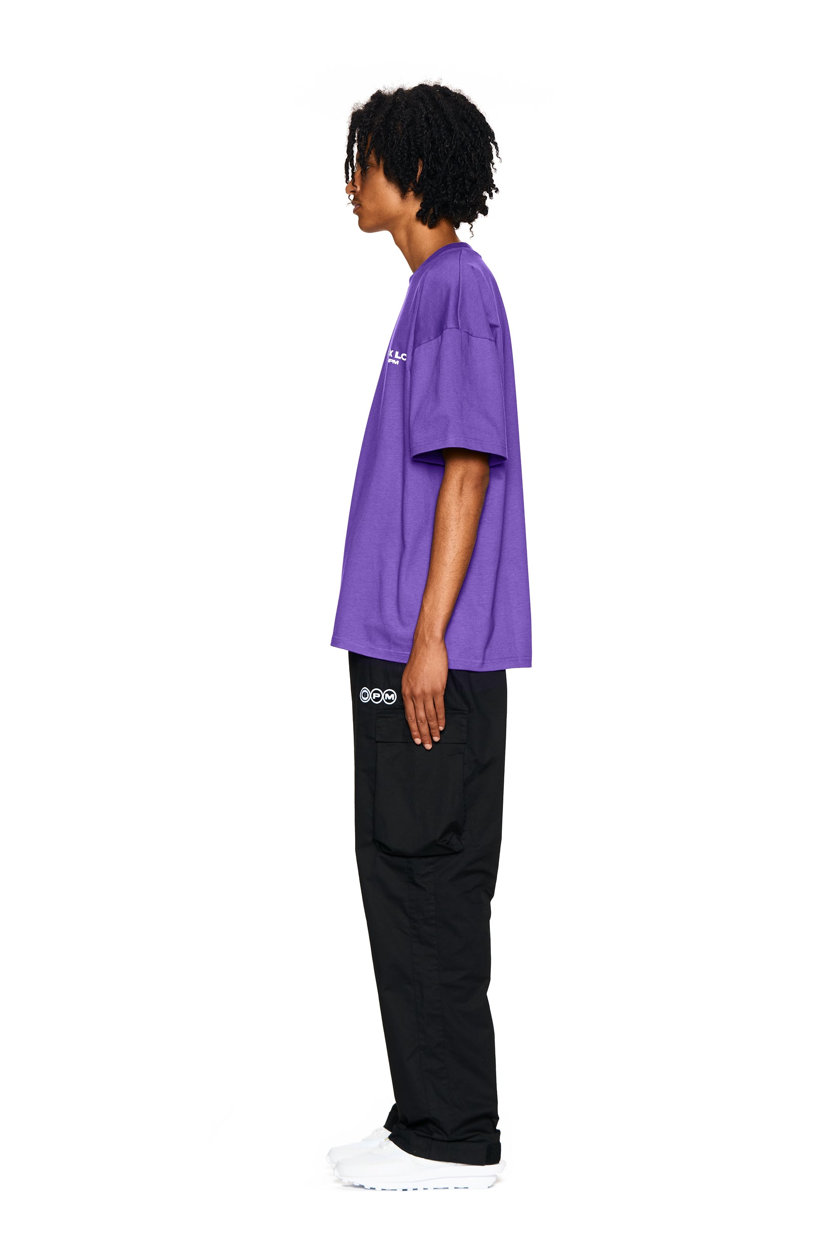 A person with medium-length curly hair stands in profile, wearing the 6PM FUCK LOVE T-SHIRT in purple, black pants with patches, and white sneakers, set against a plain white background.