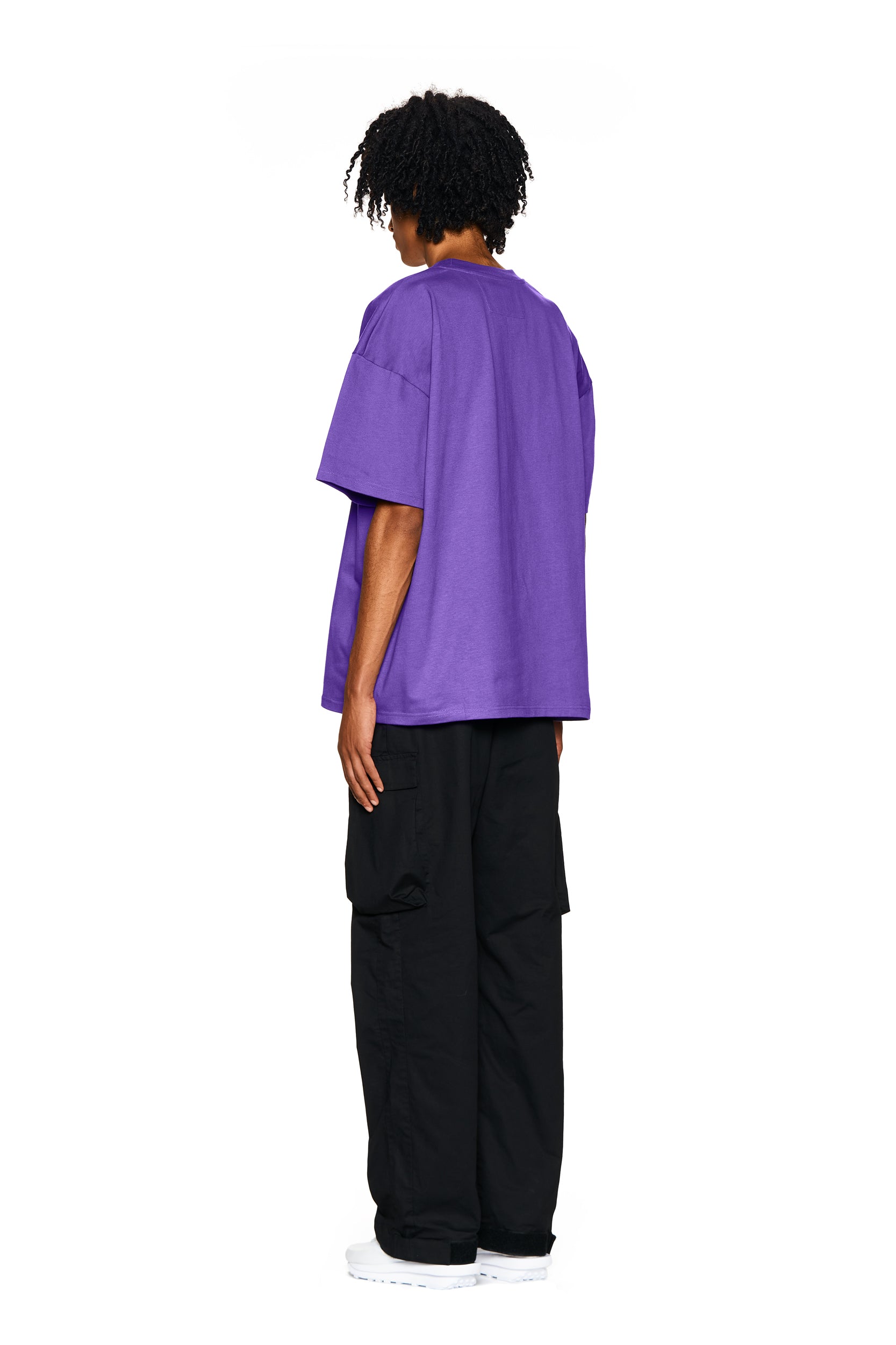 A person with curly hair stands facing away, wearing the 6PM FUCK LOVE T-SHIRT in purple, paired with black cargo pants and white sneakers, set against a plain white background.