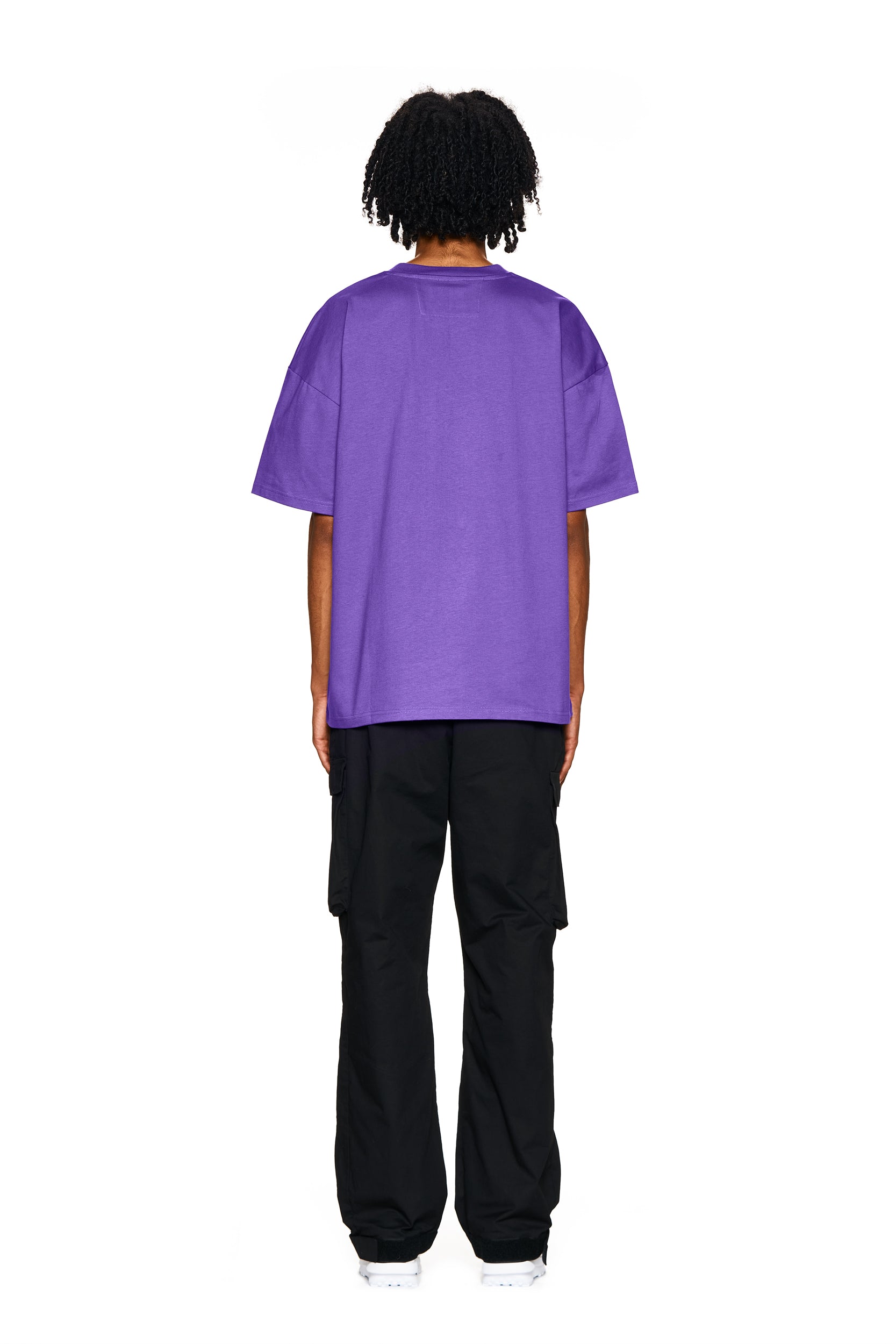 A person with curly hair faces away, wearing the 6PM FUCK LOVE T-SHIRT PURPLE, black cargo pants, and white sneakers against a plain white background.