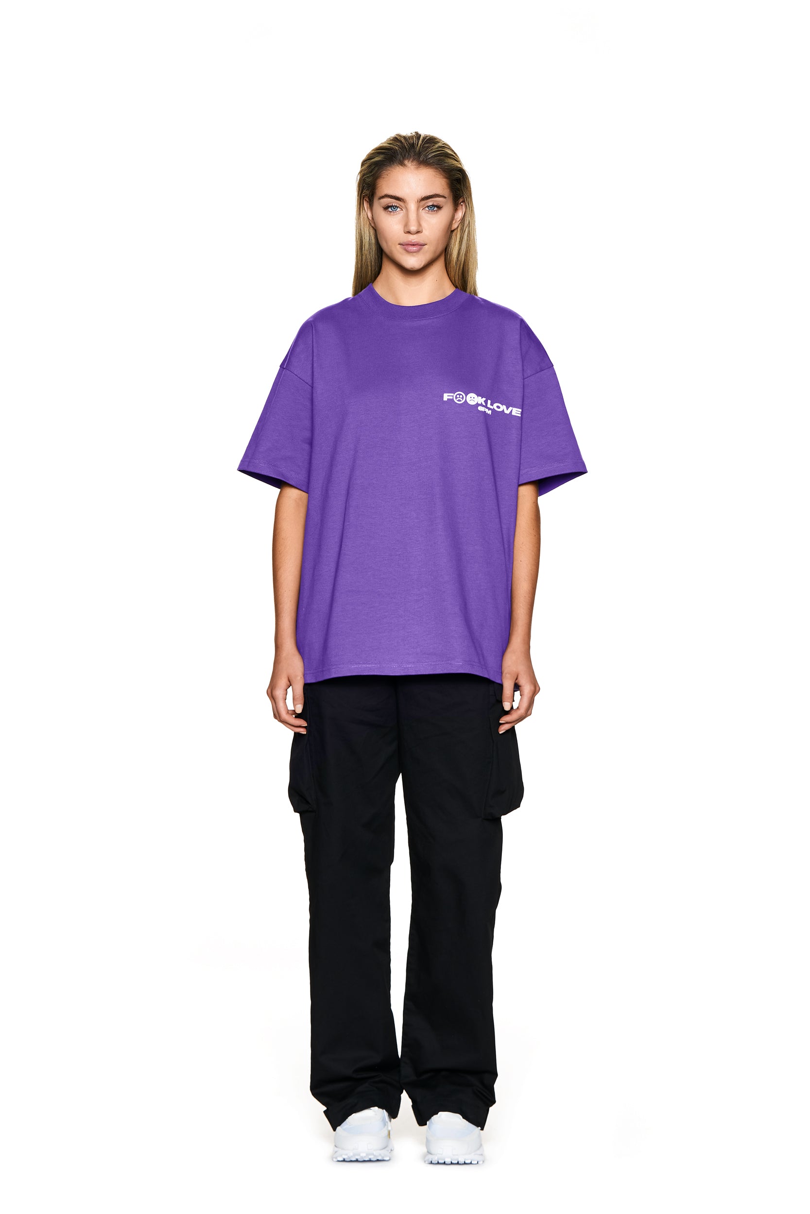 A person faces forward in a 6PM FUCK LOVE T-SHIRT PURPLE featuring a small white logo, paired with black cargo pants and white sneakers. Their straight, shoulder-length hair stands out against a plain white background.