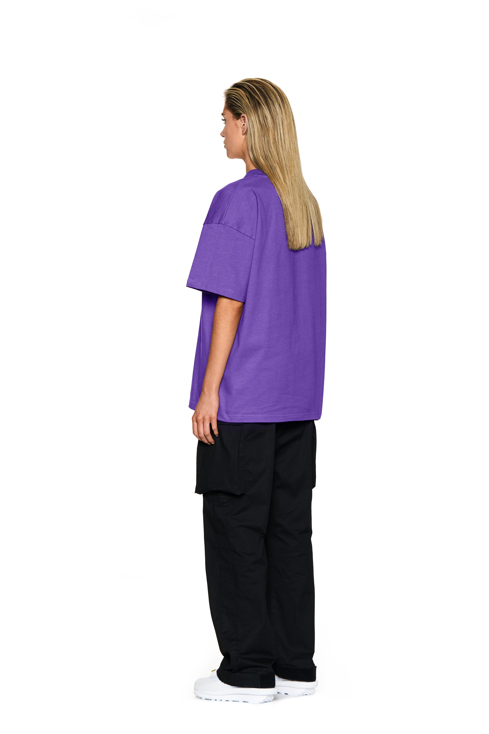 A person with straight, shoulder-length blonde hair stands facing slightly away, wearing the 6PM FUCK LOVE T-SHIRT PURPLE, black cargo pants, and white sneakers against a plain white background.