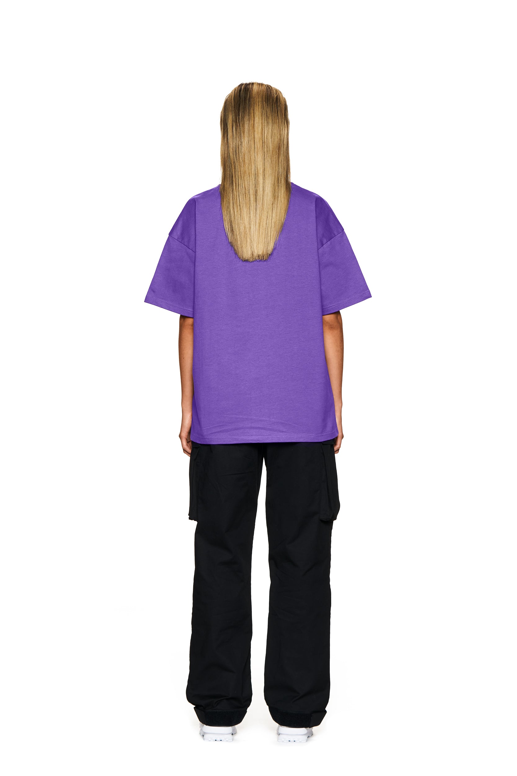 A person with long straight blonde hair wears the 6PM FUCK LOVE T-SHIRT PURPLE, black cargo pants, and white shoes, standing with their back to the camera against a plain white background.