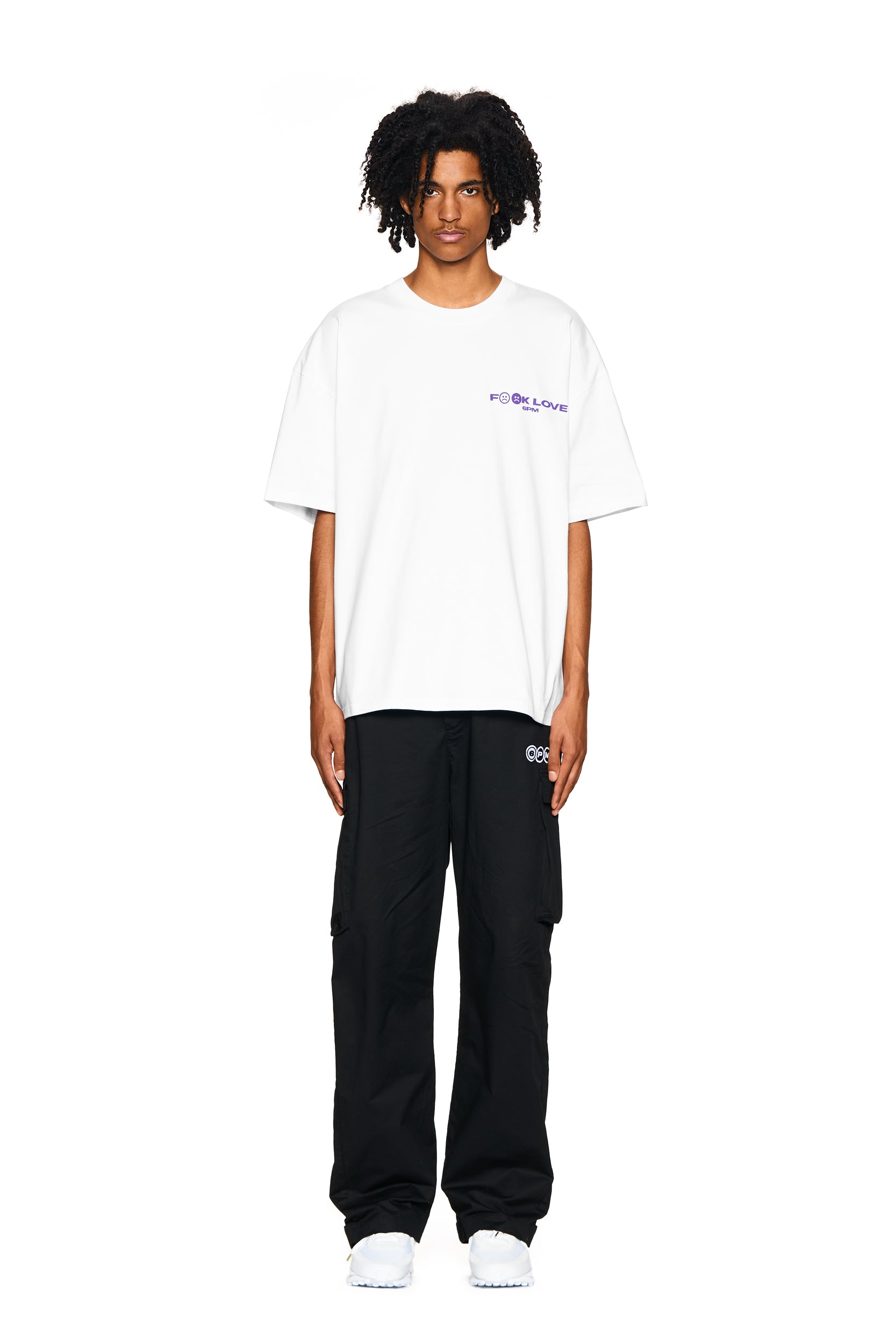 A person with curly hair faces forward, wearing the 6PM FUCK LOVE T-SHIRT WHITE with small purple text on the left chest, black cargo pants, and white sneakers, all set against a plain white background.