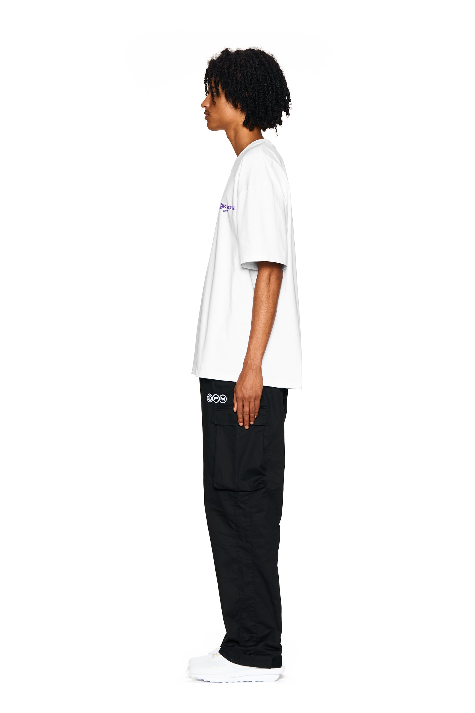 A person stands in profile against a white background, wearing the 6PM FUCK LOVE T-SHIRT WHITE, black cargo pants with a logo above the pocket, and white sneakers.