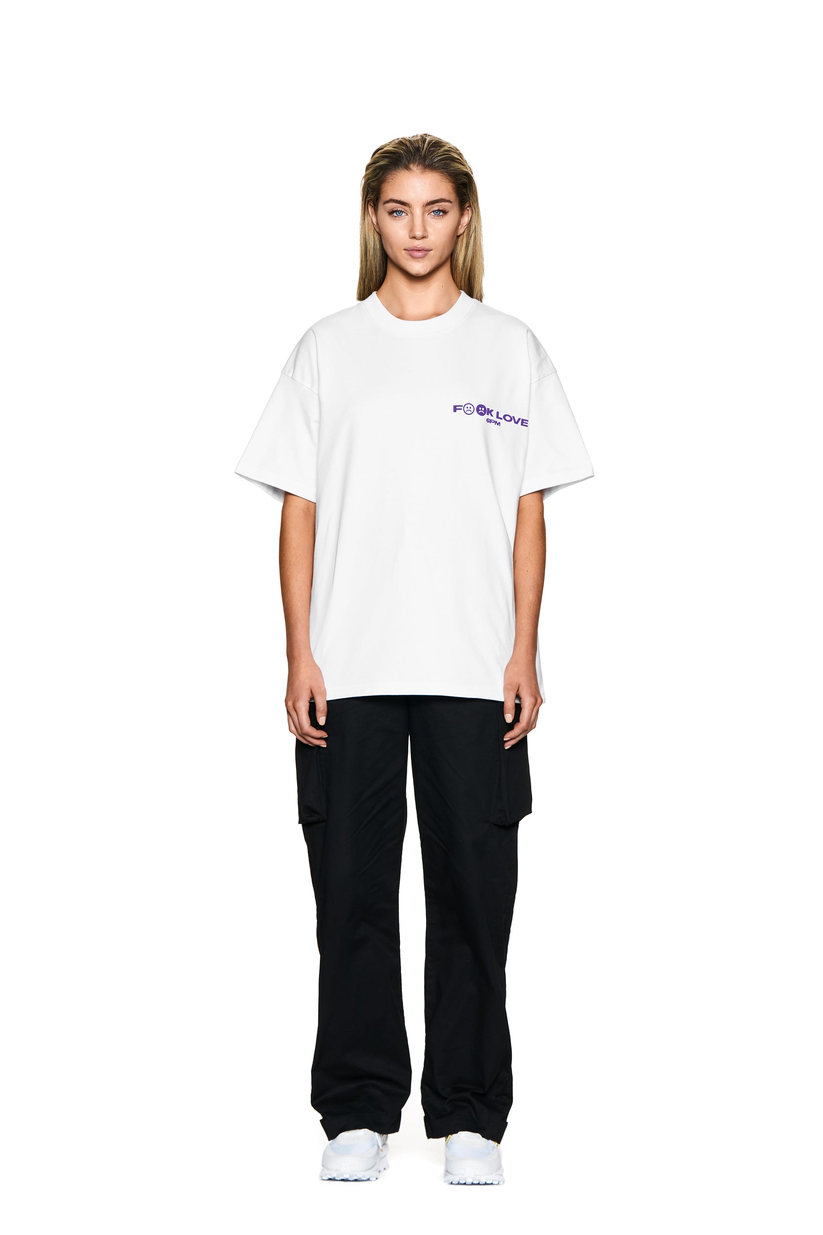 A person with long blonde hair faces forward in a 6PM FUCK LOVE T-SHIRT WHITE with purple text on the left chest, black cargo pants, and white sneakers, against a plain white background.