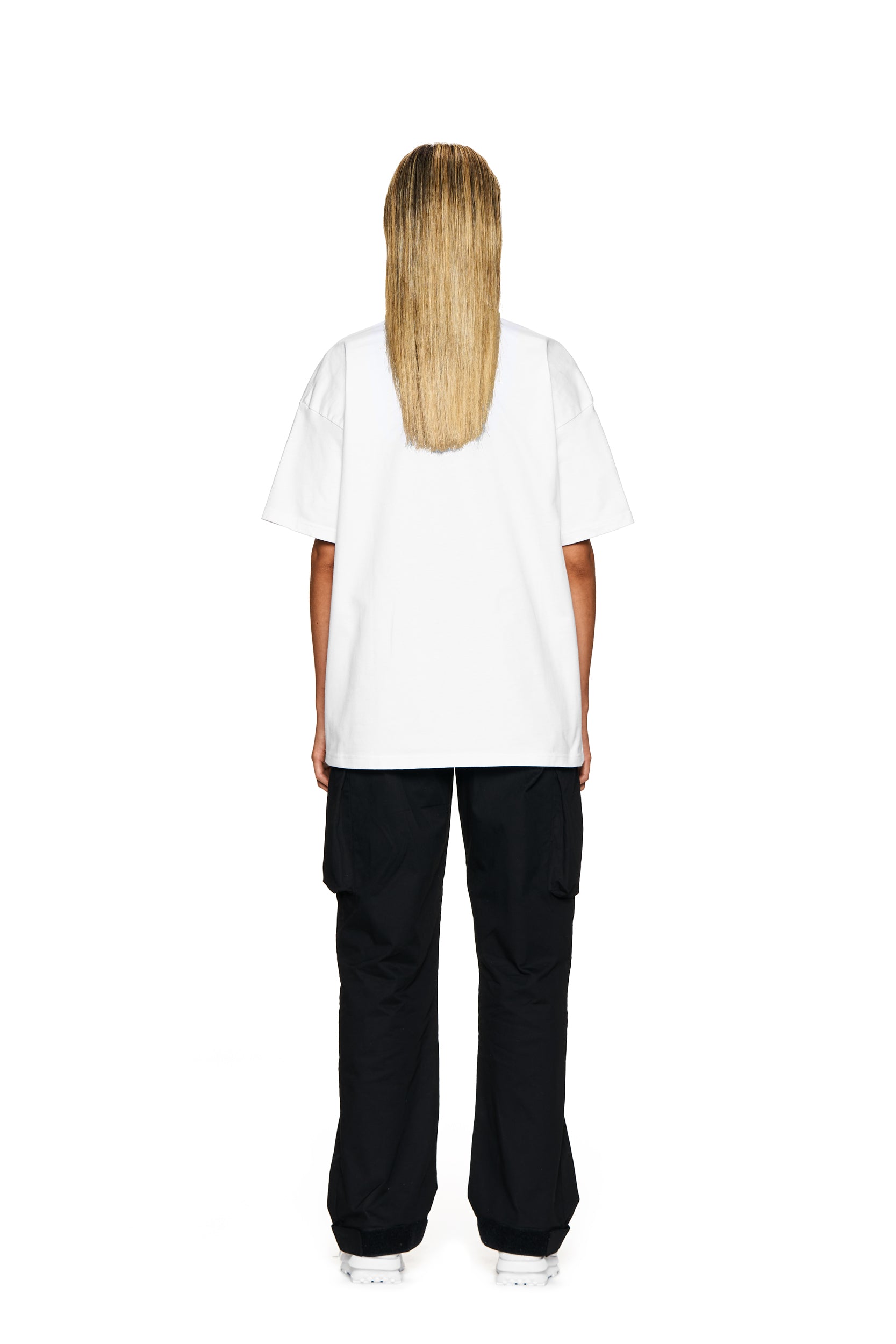 A person with long blonde hair faces away, wearing the 6PM FUCK LOVE T-SHIRT WHITE, loose black cargo pants, and white sneakers against a white background.
