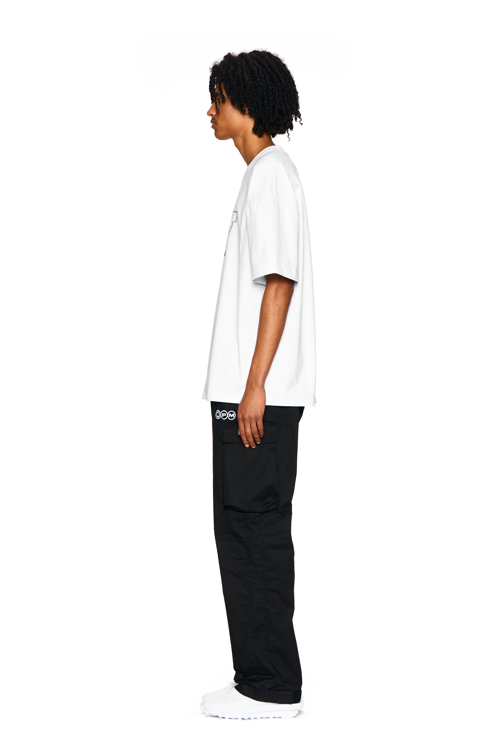 A person with curly hair stands in profile facing left, wearing the 6PM BLING T-SHIRT WHITE, black cargo pants with a small logo, and white sneakers, against a plain white background.