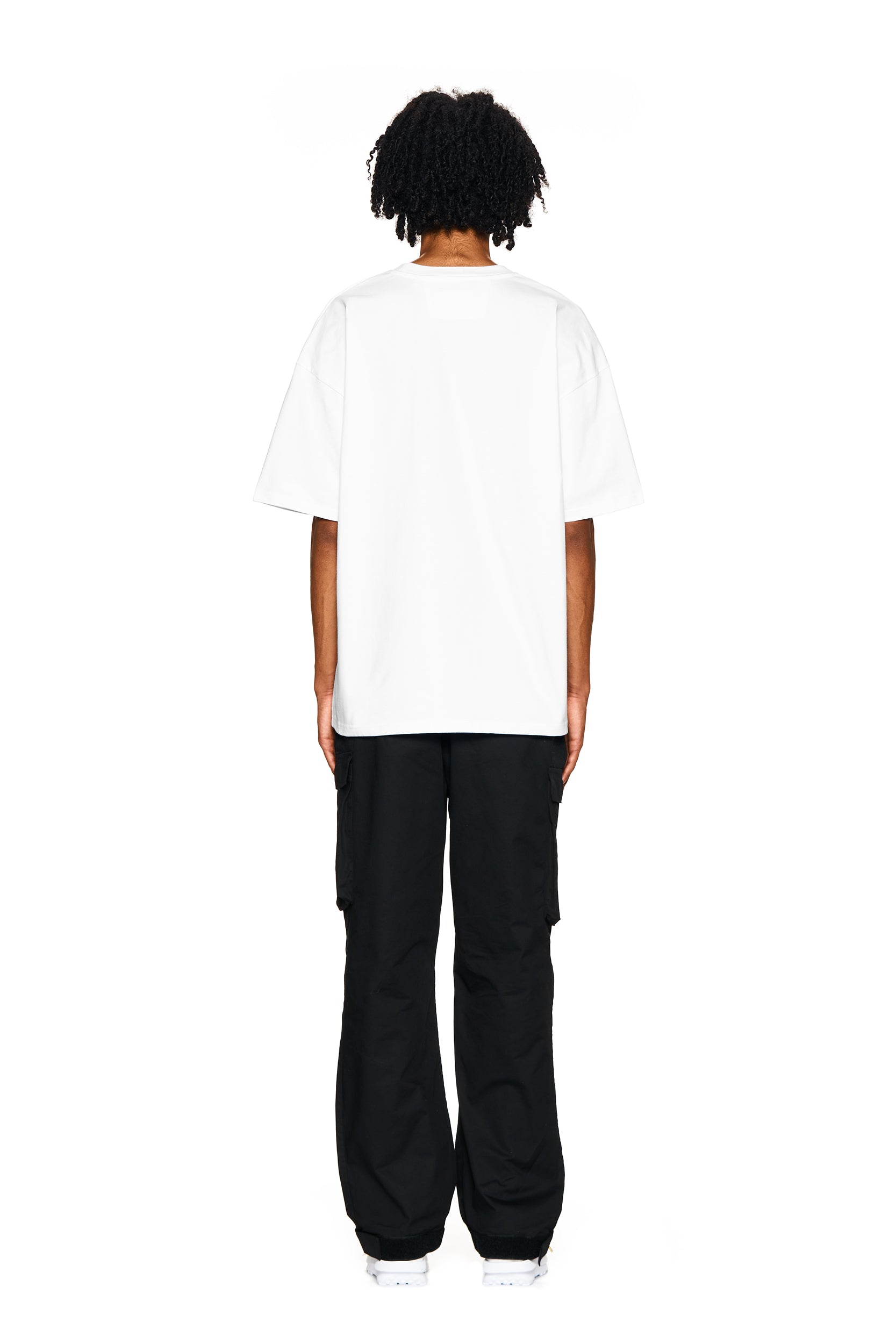 A person with short curly hair stands facing away, wearing the 6PM BLING T-SHIRT WHITE, loose black pants, and white sneakers, against a white background.