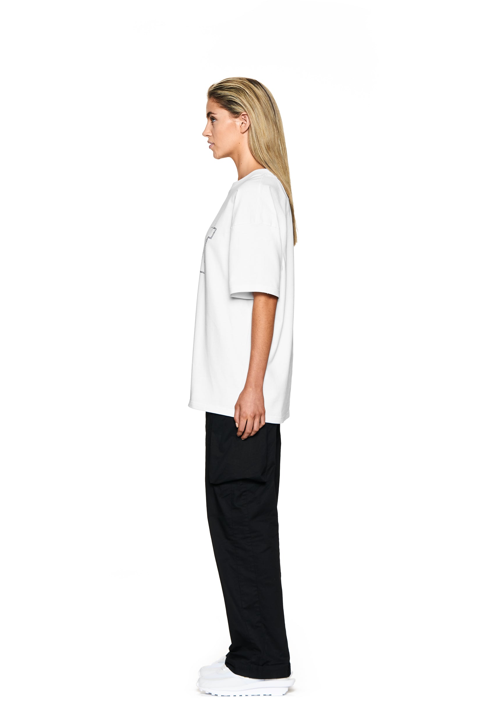 A person with straight, shoulder-length blonde hair stands in profile, wearing the 6PM BLING T-SHIRT WHITE, black pants, and white sneakers against a plain white background.