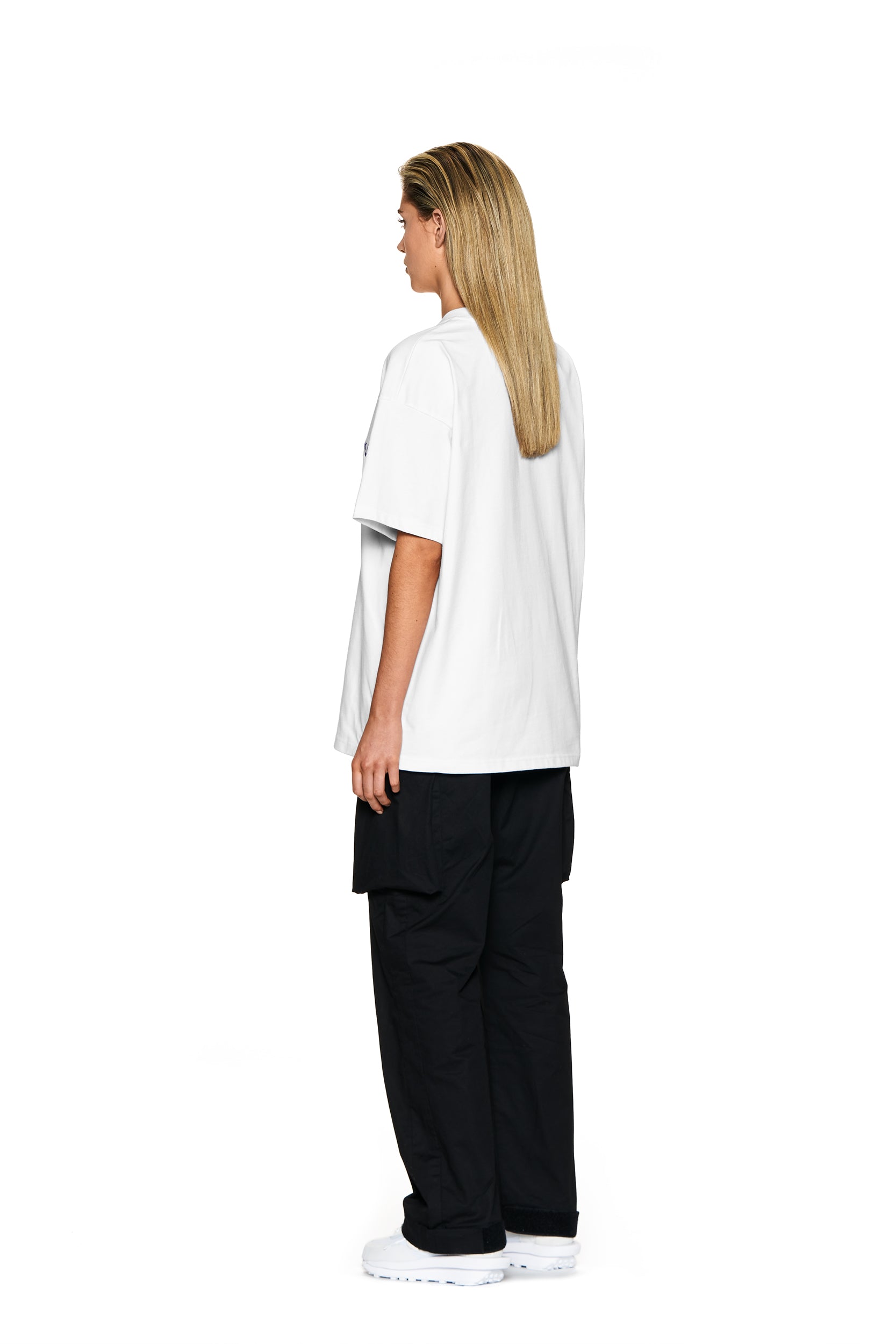 A person with straight, shoulder-length blonde hair stands sideways against a white background, wearing the 6PM BLING T-SHIRT WHITE, black cargo pants, and white shoes.