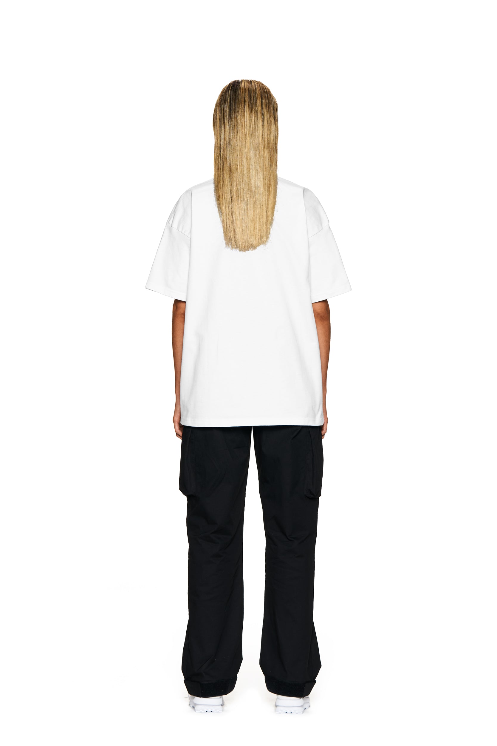 A person with long, straight blonde hair stands facing away, wearing the 6PM BLING T-SHIRT WHITE, black cargo pants, and white shoes against a white background.