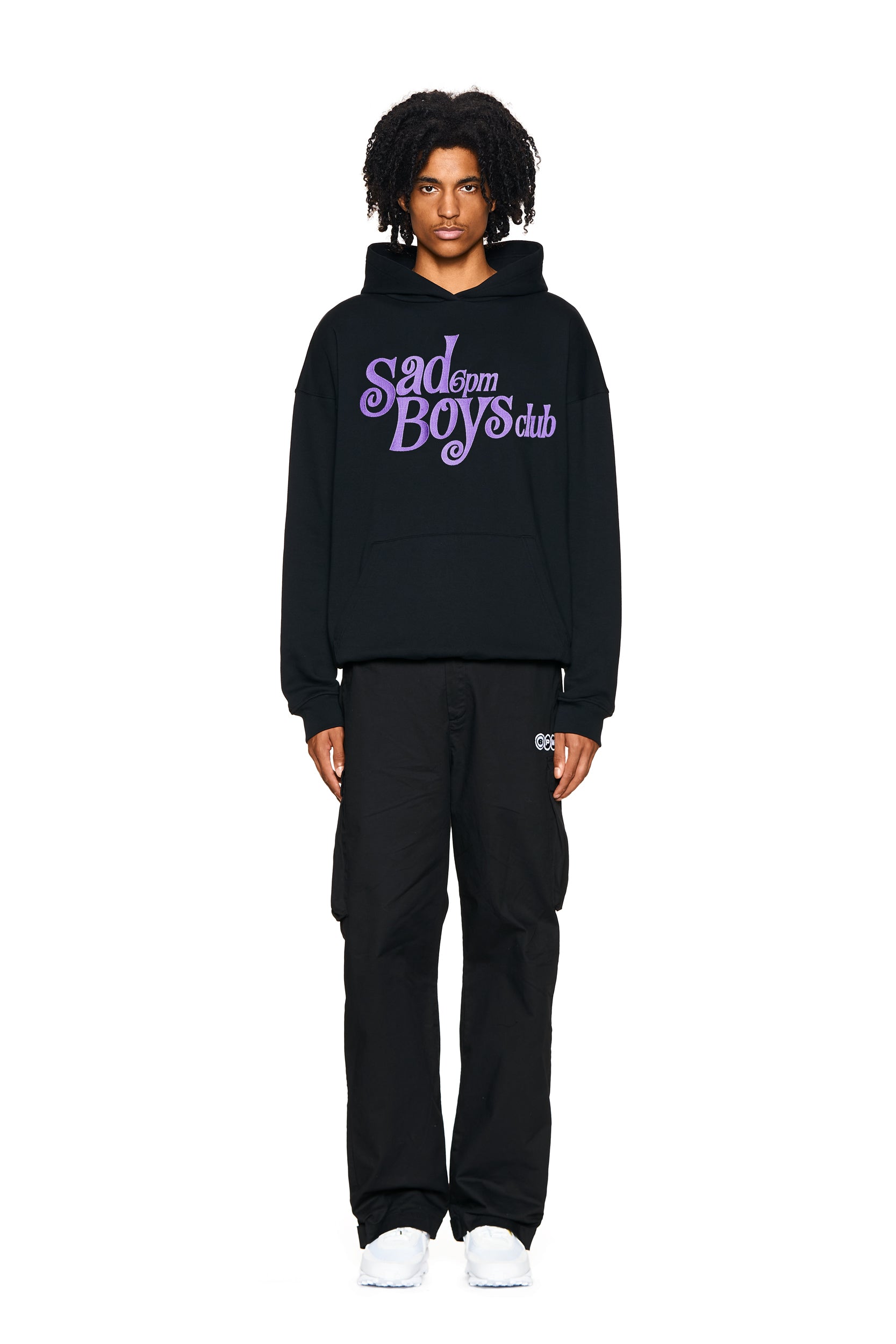A person stands facing forward against a plain white background, wearing the 6PM SAD BOYS HOODIE BLACK, black pants, and white sneakers. The hoodie features "Sad Boys Club" written in purple and white letters.