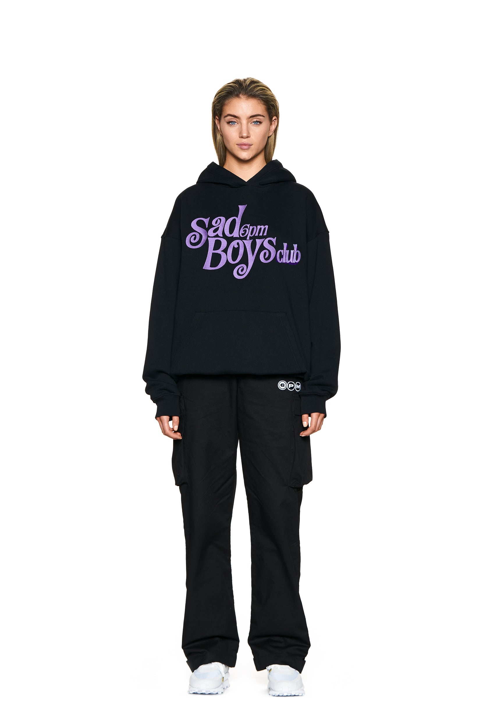 A person faces forward against a plain white background, wearing the 6PM SAD BOYS HOODIE BLACK with purple text, paired with black cargo pants and white sneakers.