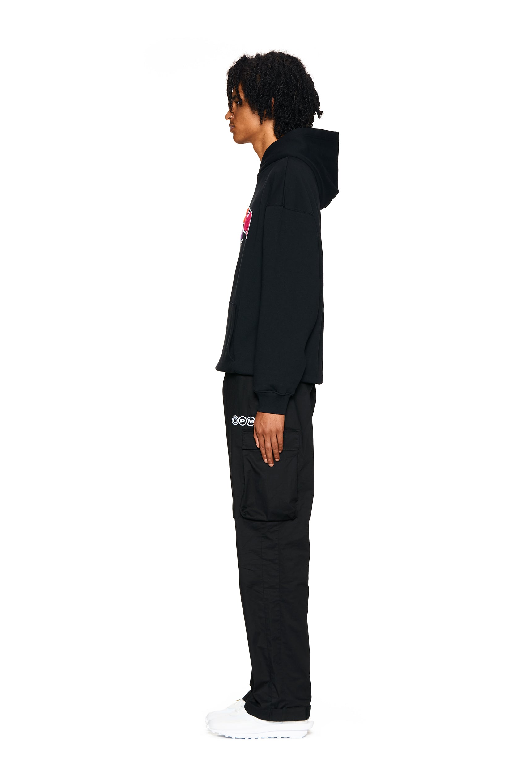 A person with curly hair stands in profile against a white background, wearing the 6PM LOGO HOODIE BLACK, black cargo pants with logo pockets, and white sneakers.