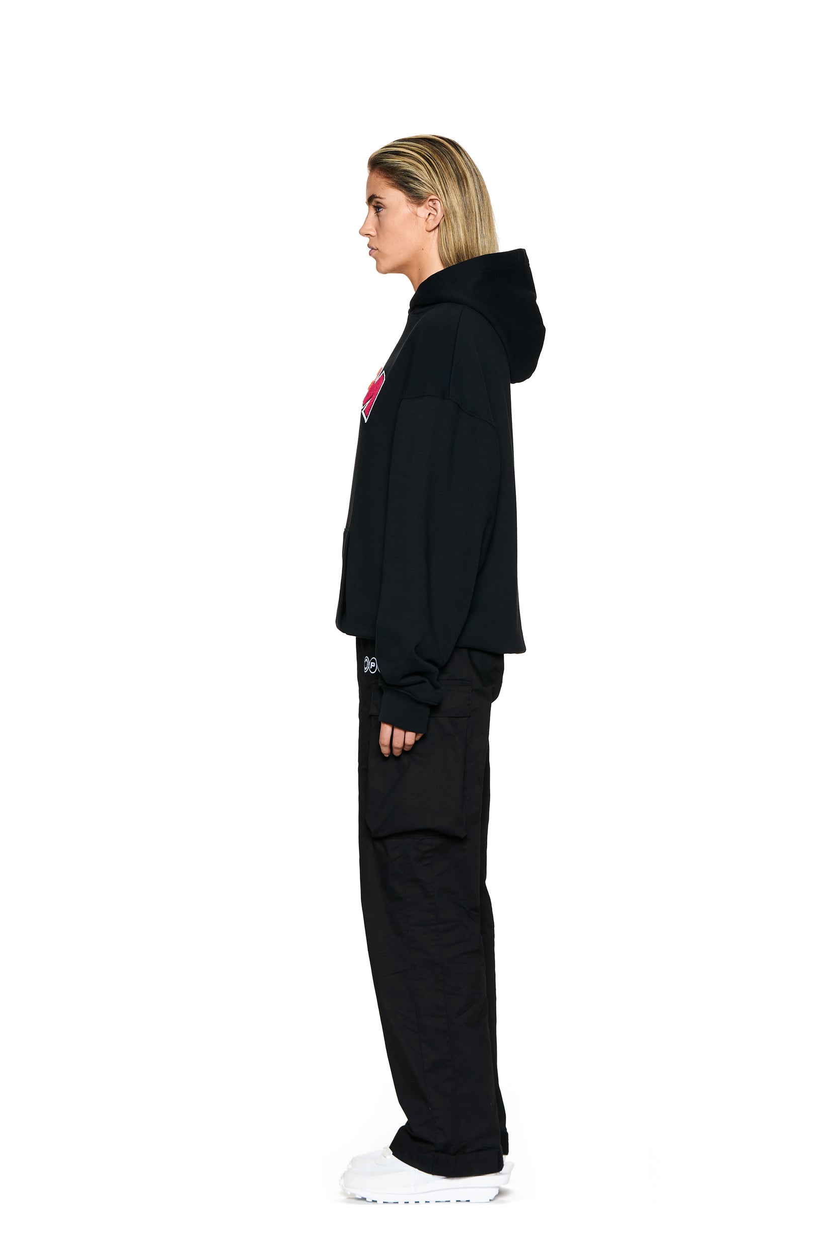 A person with blonde hair stands in profile, facing left, wearing the 6PM LOGO HOODIE BLACK, black pants, and white sneakers against a plain white background.