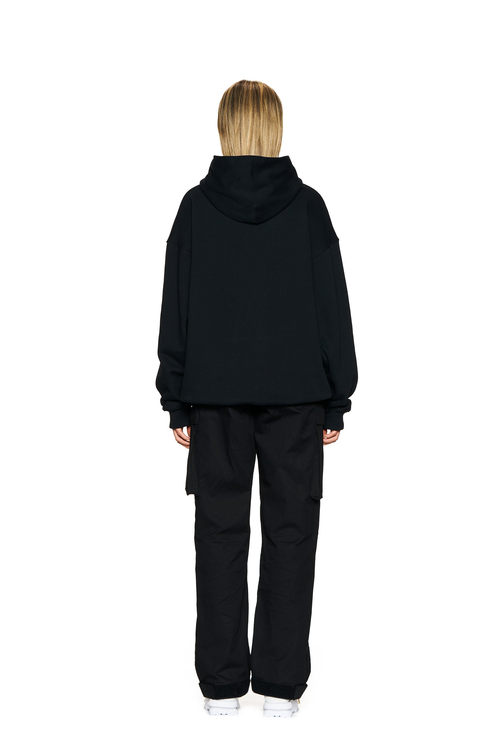 A person with shoulder-length blonde hair faces away, wearing the 6PM LOGO HOODIE BLACK paired with black pants and white shoes against a plain white background.