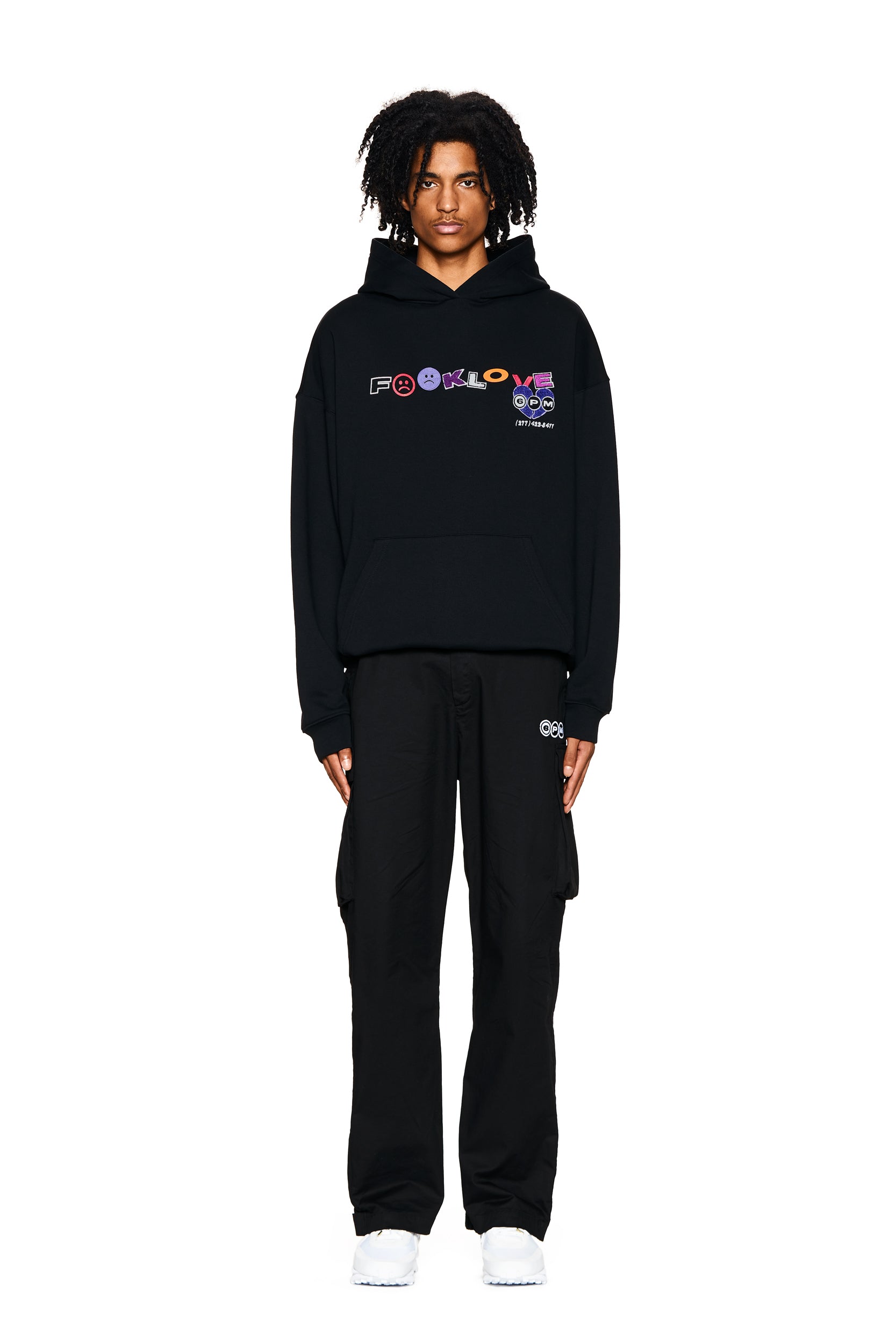 A person with short, curly hair faces forward wearing the 6PM FUCK LOVE HOODIE BLACK featuring colorful text and symbols, paired with black cargo pants and white sneakers, against a plain white background.