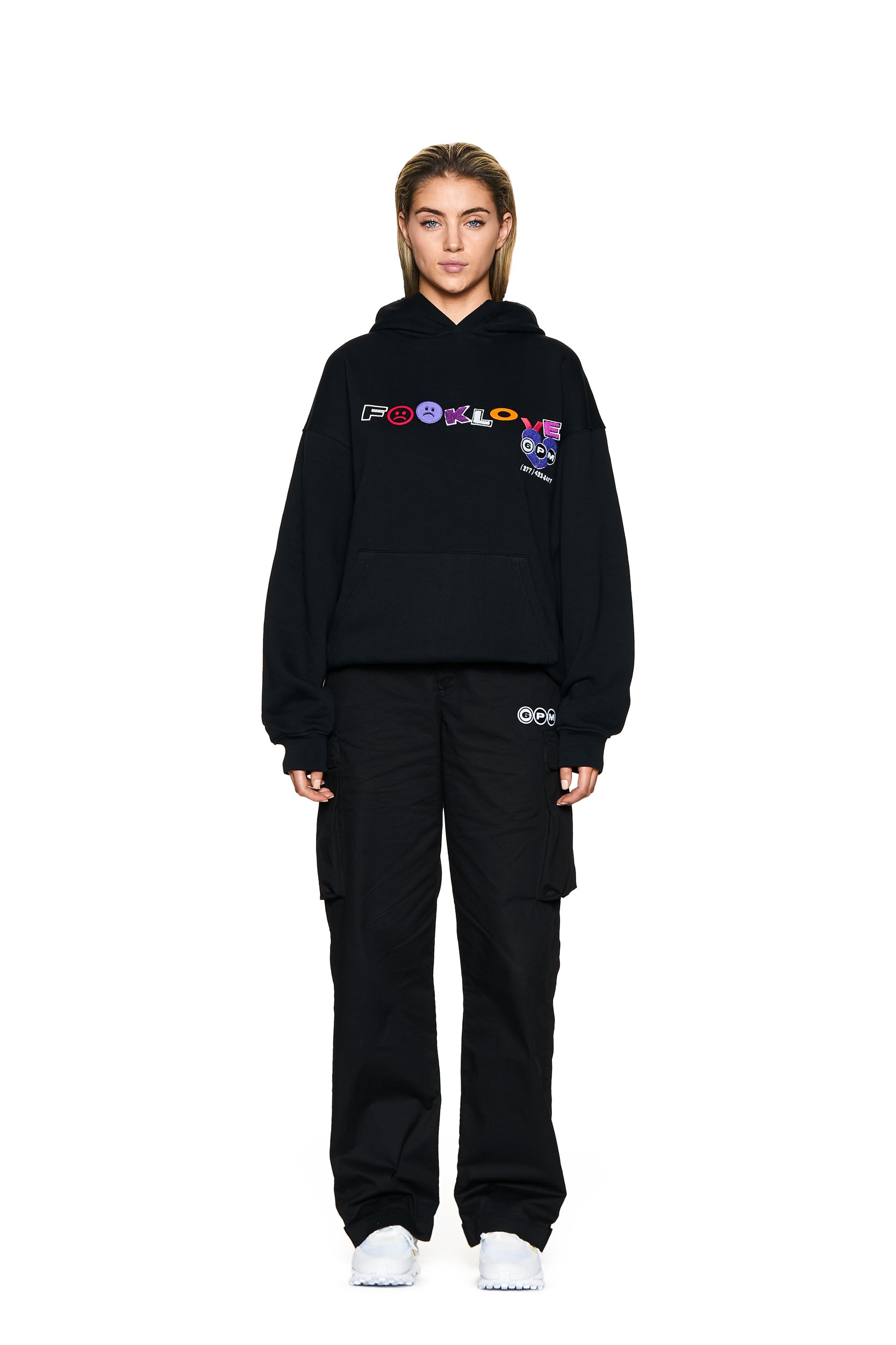 A person stands facing forward against a plain white background, wearing the 6PM FUCK LOVE HOODIE BLACK with colorful text and graphics on the front, paired with black cargo pants and white sneakers.
