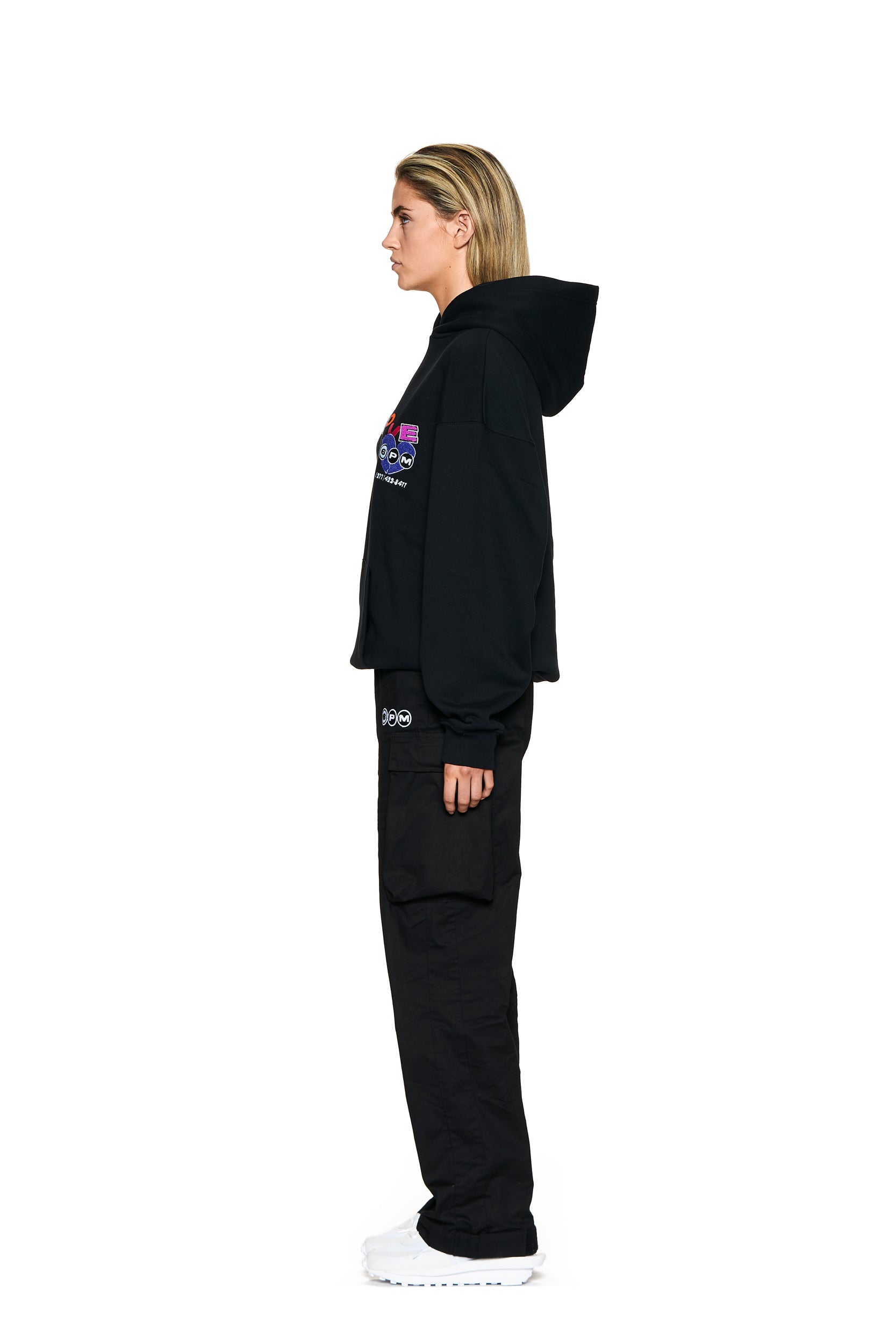 A woman with blonde hair stands in profile against a plain white background, wearing 6PM's CARGO PANTS BLACK, a black hoodie with colorful designs, and white sneakers.