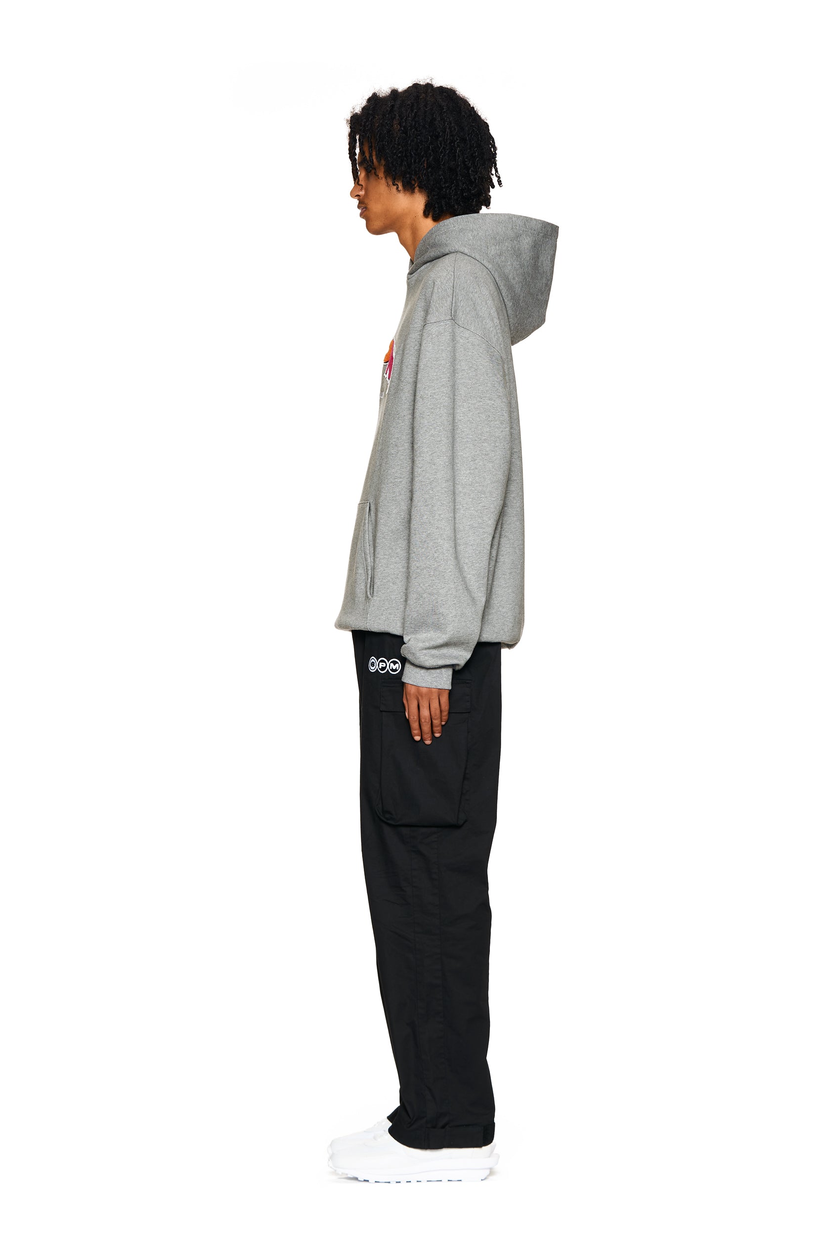 A person with curly hair stands in profile against a white background, wearing the 6PM LOGO HOODIE GREY MELANGE, black pants, and white shoes.