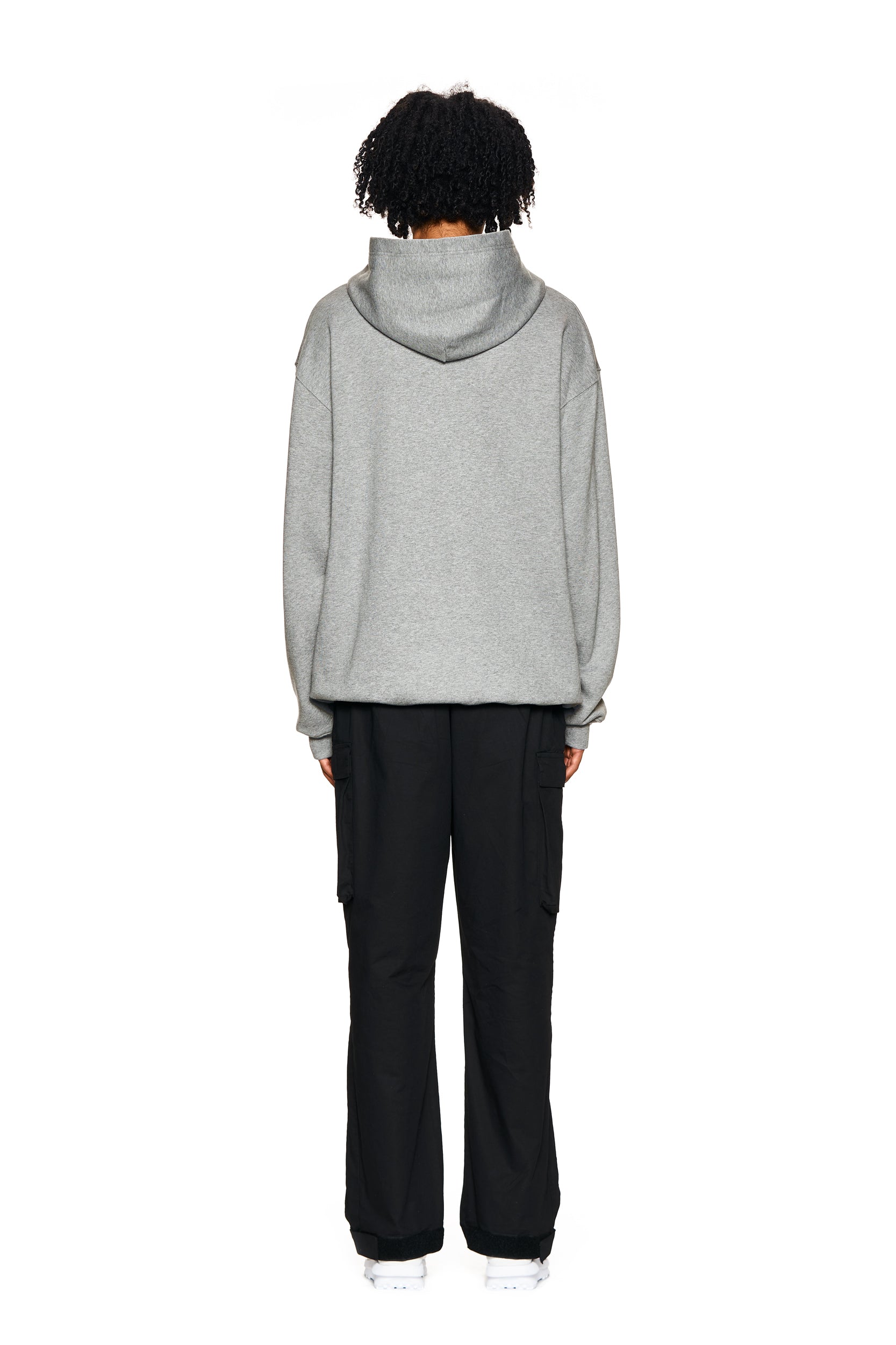 A person with curly hair stands facing away, wearing the 6PM LOGO HOODIE GREY MELANGE, paired with black cargo pants and white shoes, against a plain white background.