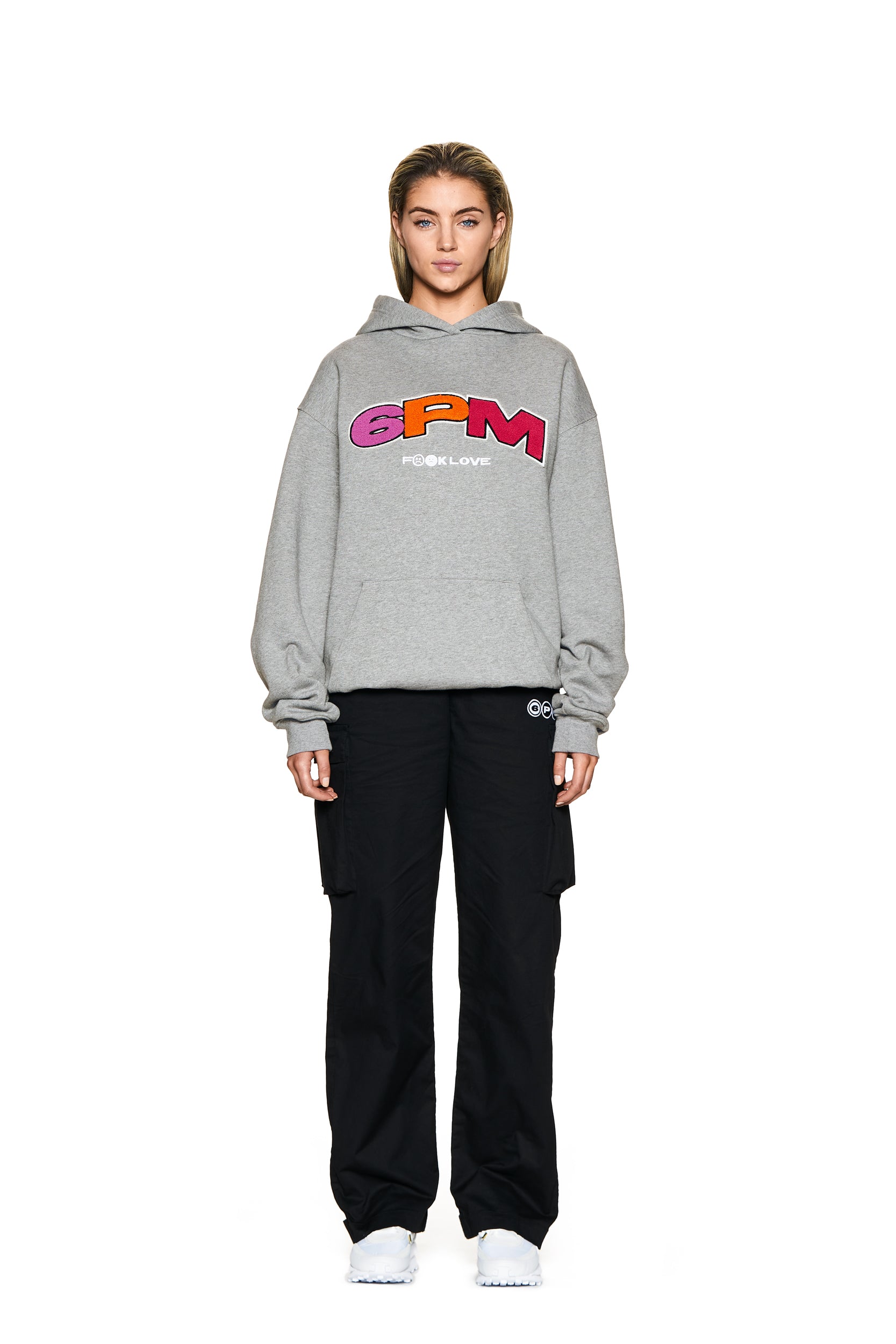 A person faces forward on a plain white background, wearing the 6PM LOGO HOODIE GREY MELANGE with colorful “6PM” text and “PEACE & LOVE” below, paired with black cargo pants and white sneakers.