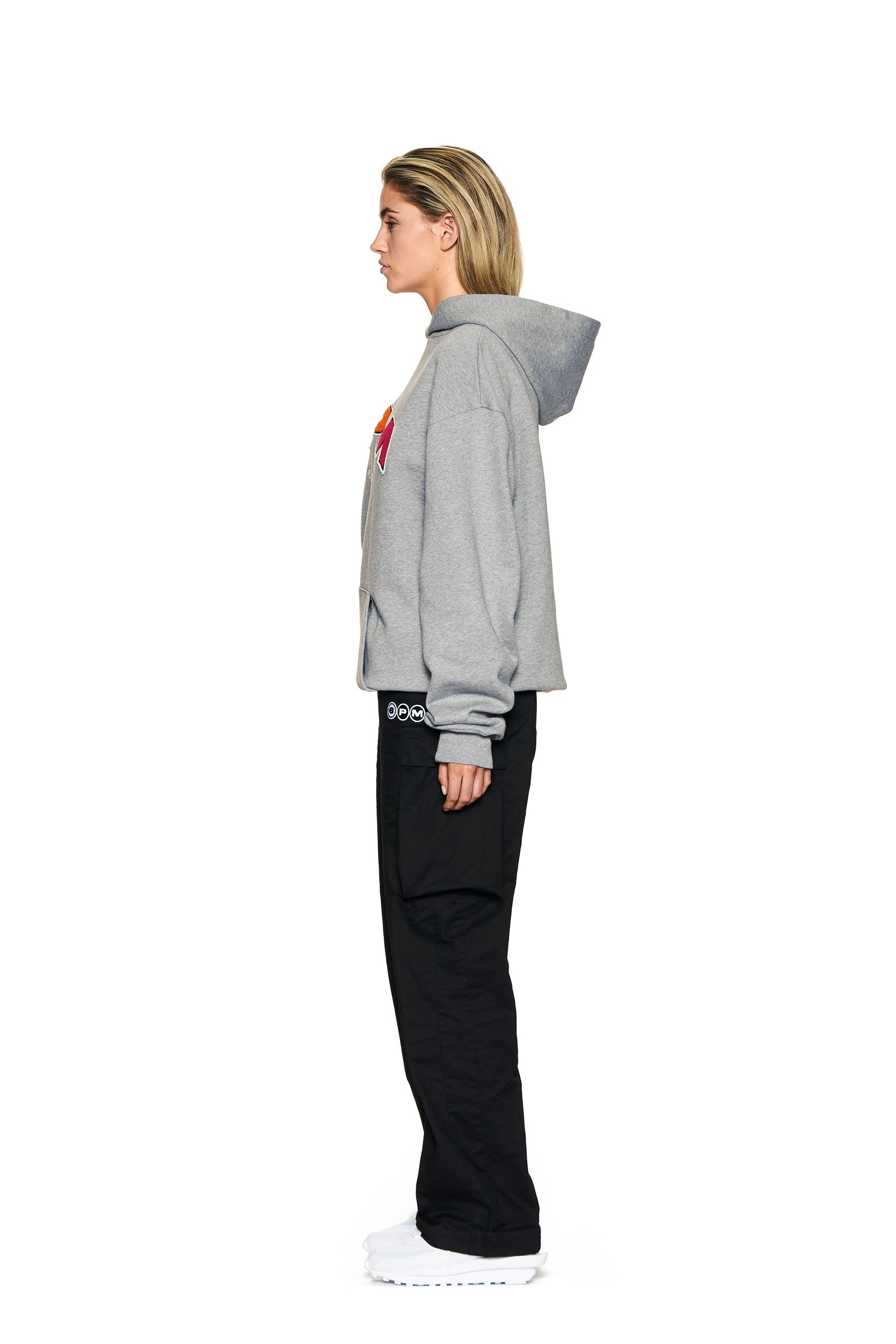 A person in profile faces left, wearing the 6PM LOGO HOODIE GREY MELANGE, black pants, and white shoes against a plain white background.