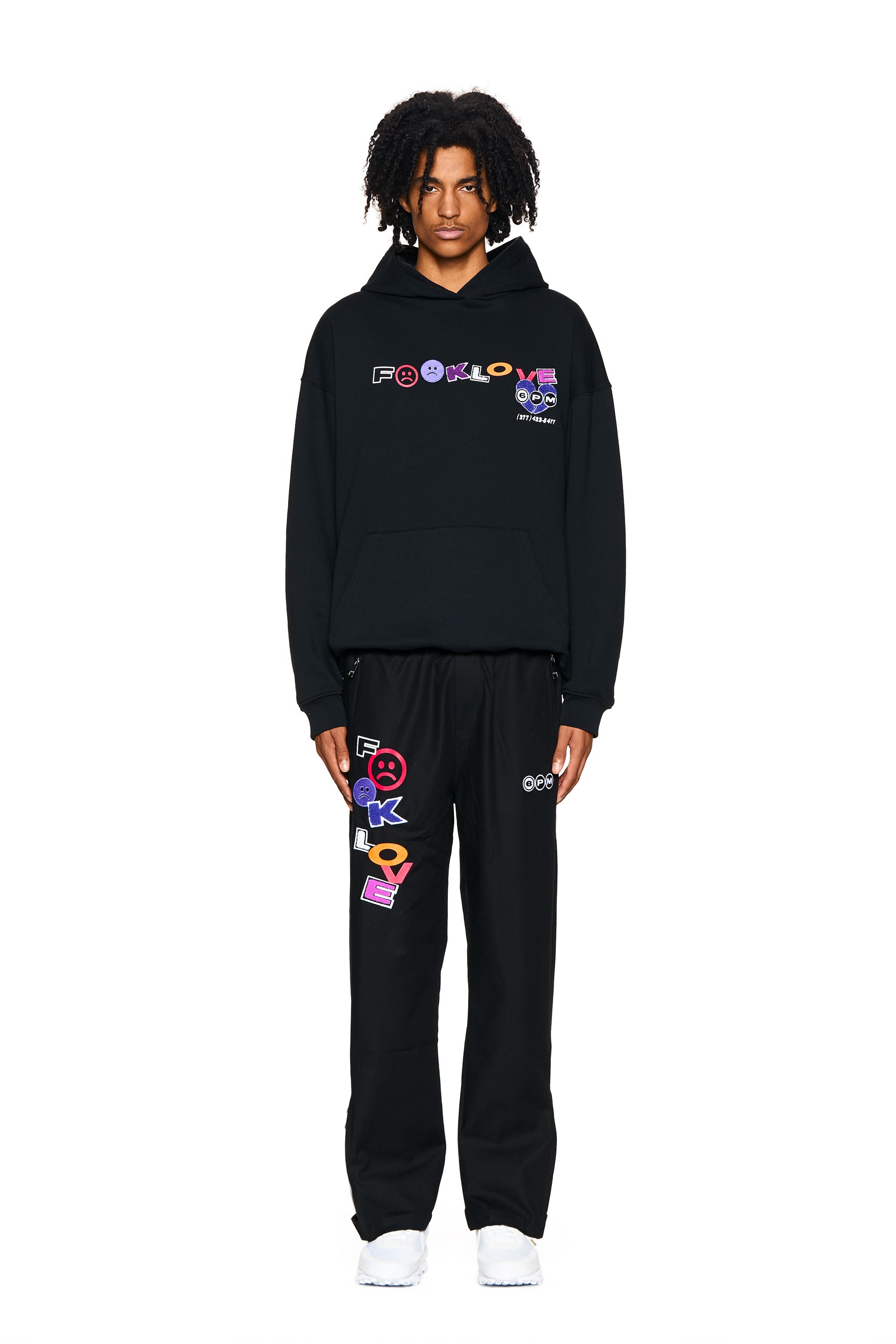 A person faces forward in a black hoodie and 6PM FUCK LOVE TRACKPANTS BLACK, featuring bold, colorful graphics and text. They have medium-length curly hair, white sneakers, and stand against a plain white background.