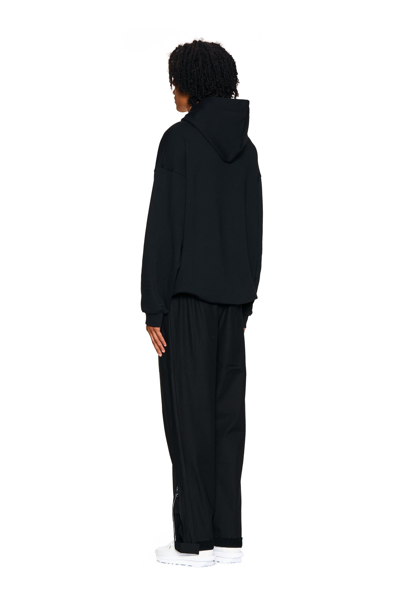 A person with curly hair, wearing 6PM's FUCK LOVE TRACKPANTS BLACK and a black hoodie, stands facing away from the camera against a plain white background.