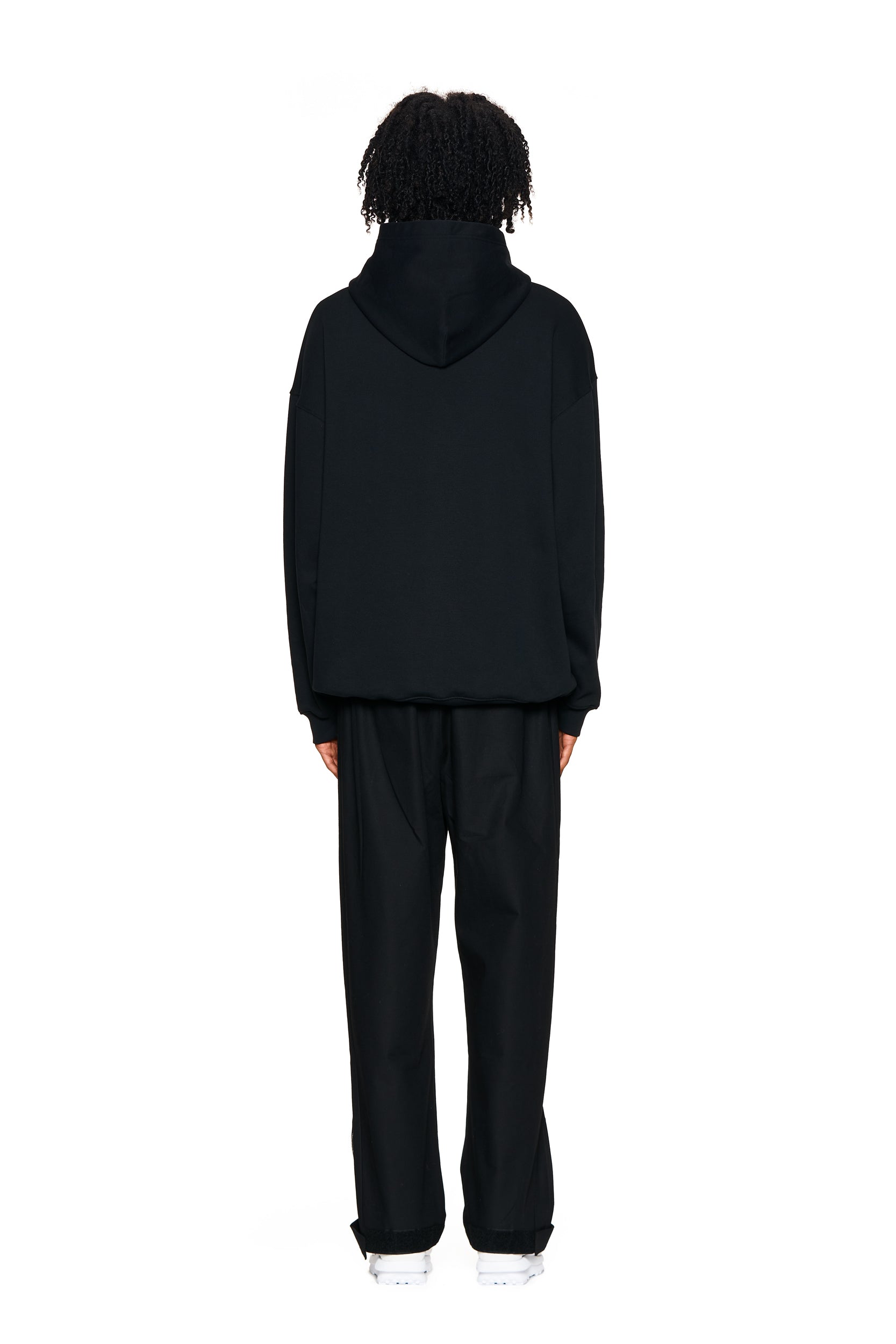 A person faces away from the camera against a plain white background, wearing 6PM's FUCK LOVE TRACKPANTS in black paired with a black hoodie and white shoes.