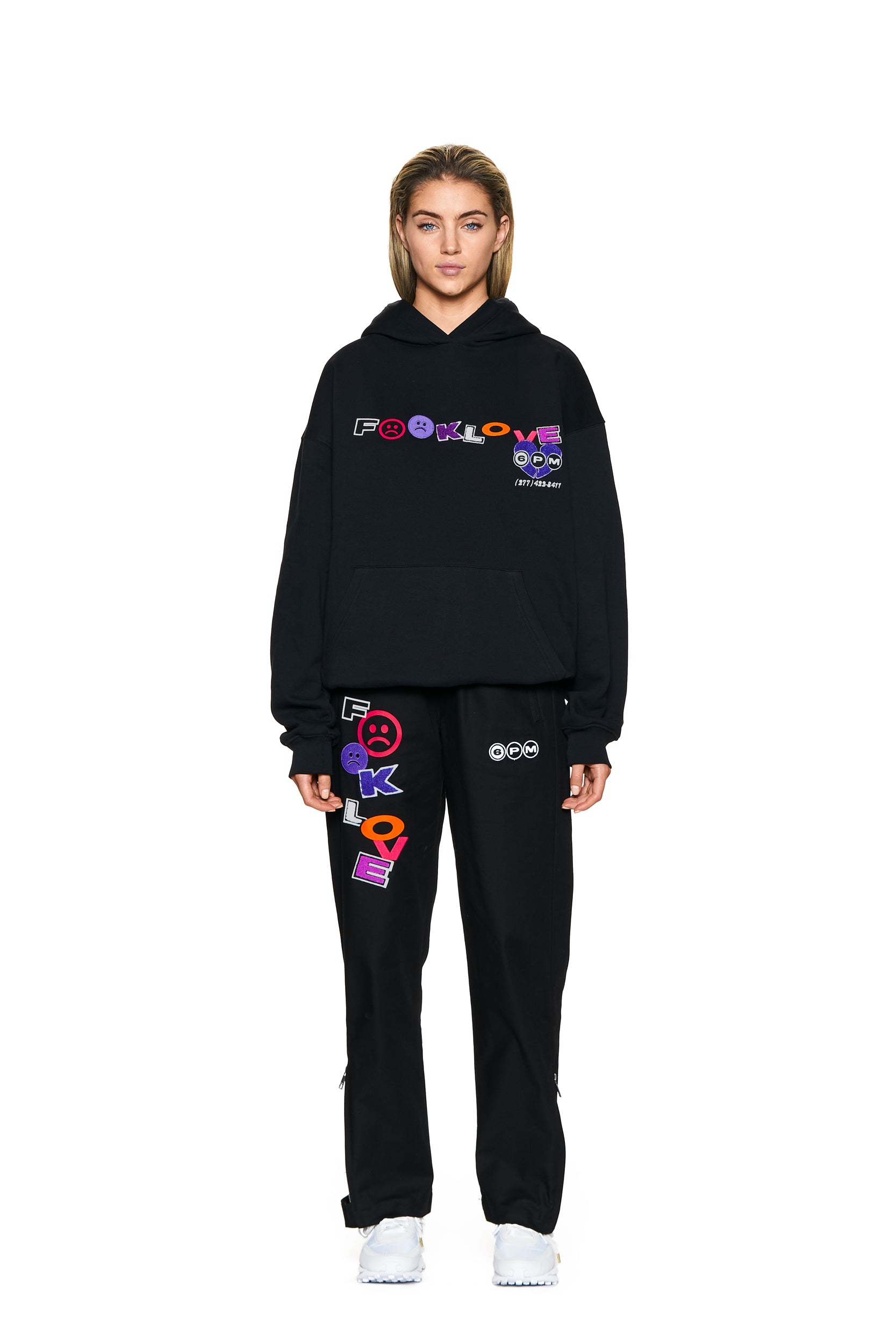 A person faces forward on a plain white background, wearing 6PM’s black FUCK LOVE TRACKPANTS with colorful text and graphic designs, a black hoodie, white sneakers, and has light brown hair.