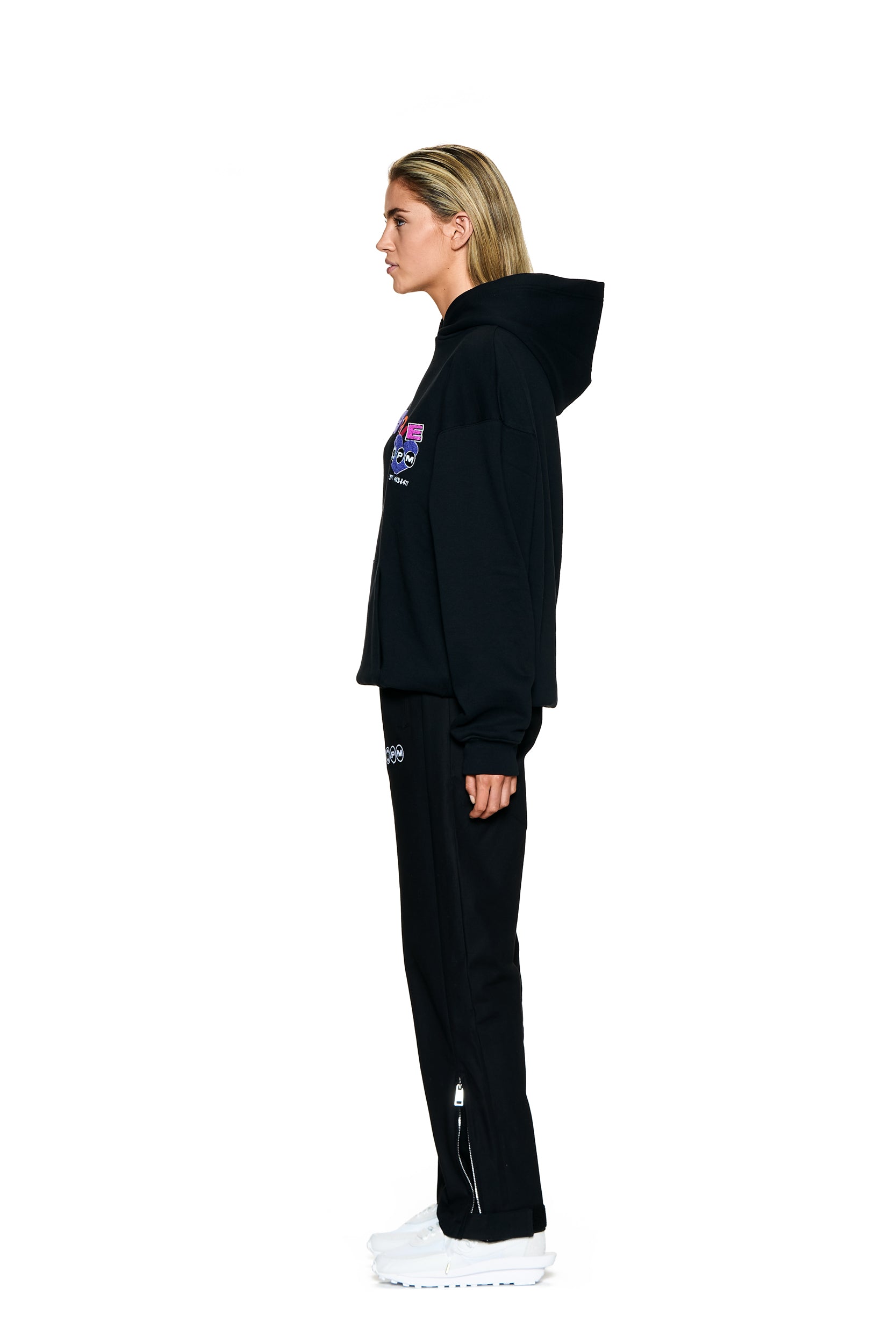 A person with blonde hair stands in profile against a white background, wearing a black hoodie, 6PM FUCK LOVE TRACKPANTS BLACK, and white sneakers.