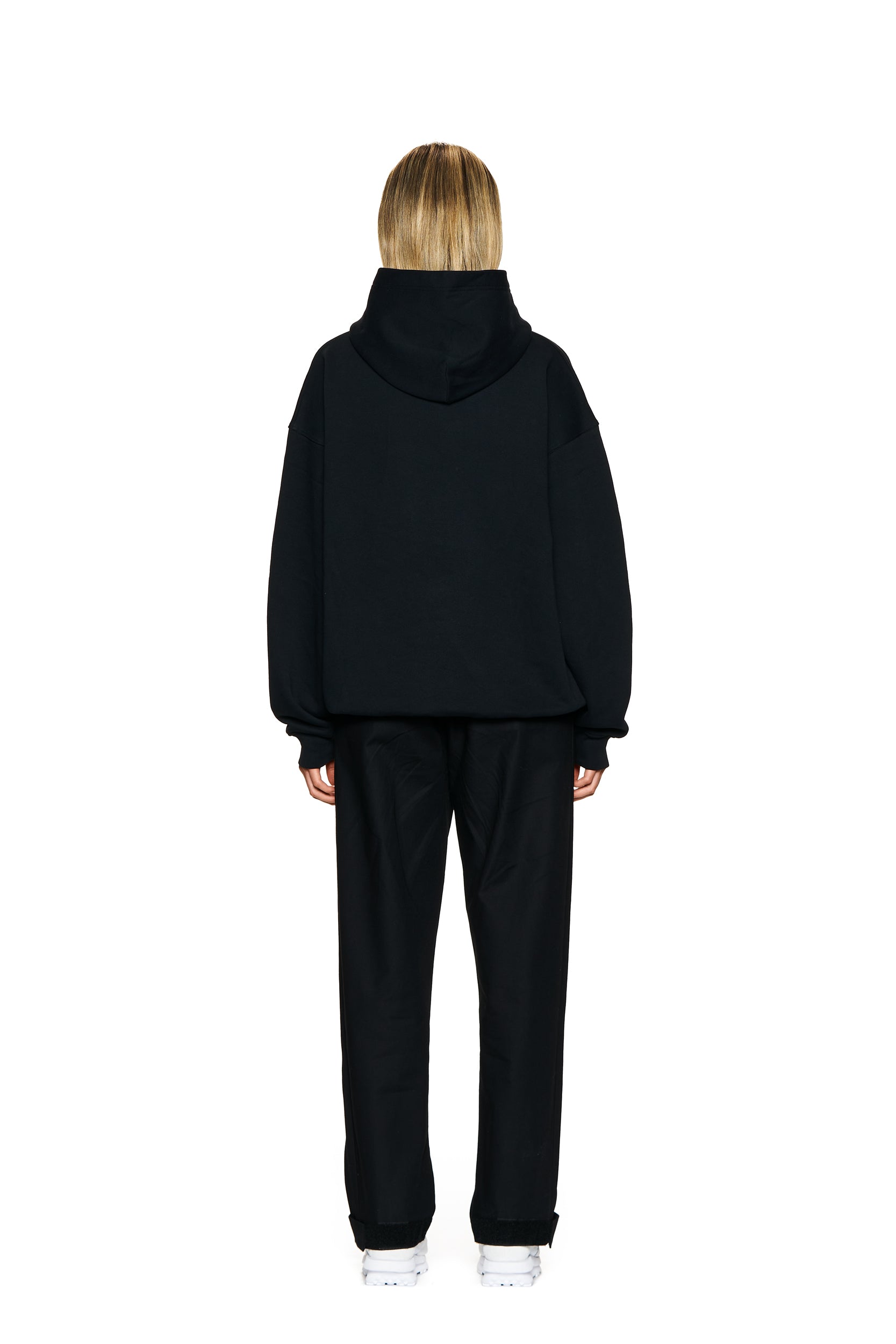 A person with blonde hair, dressed in a black hoodie, 6PM FUCK LOVE TRACKPANTS BLACK, and white sneakers stands facing away from the camera against a plain white background.