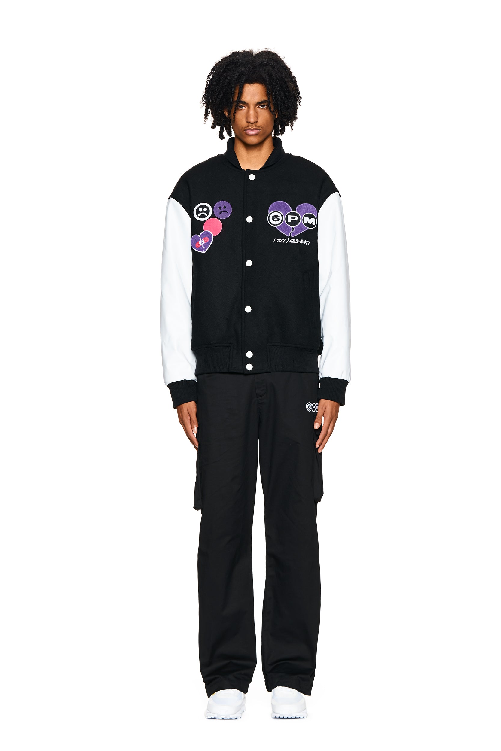 A person faces forward against a white background wearing the 6PM COLLEGE JACKET BLACK styled with black cargo pants and white sneakers.