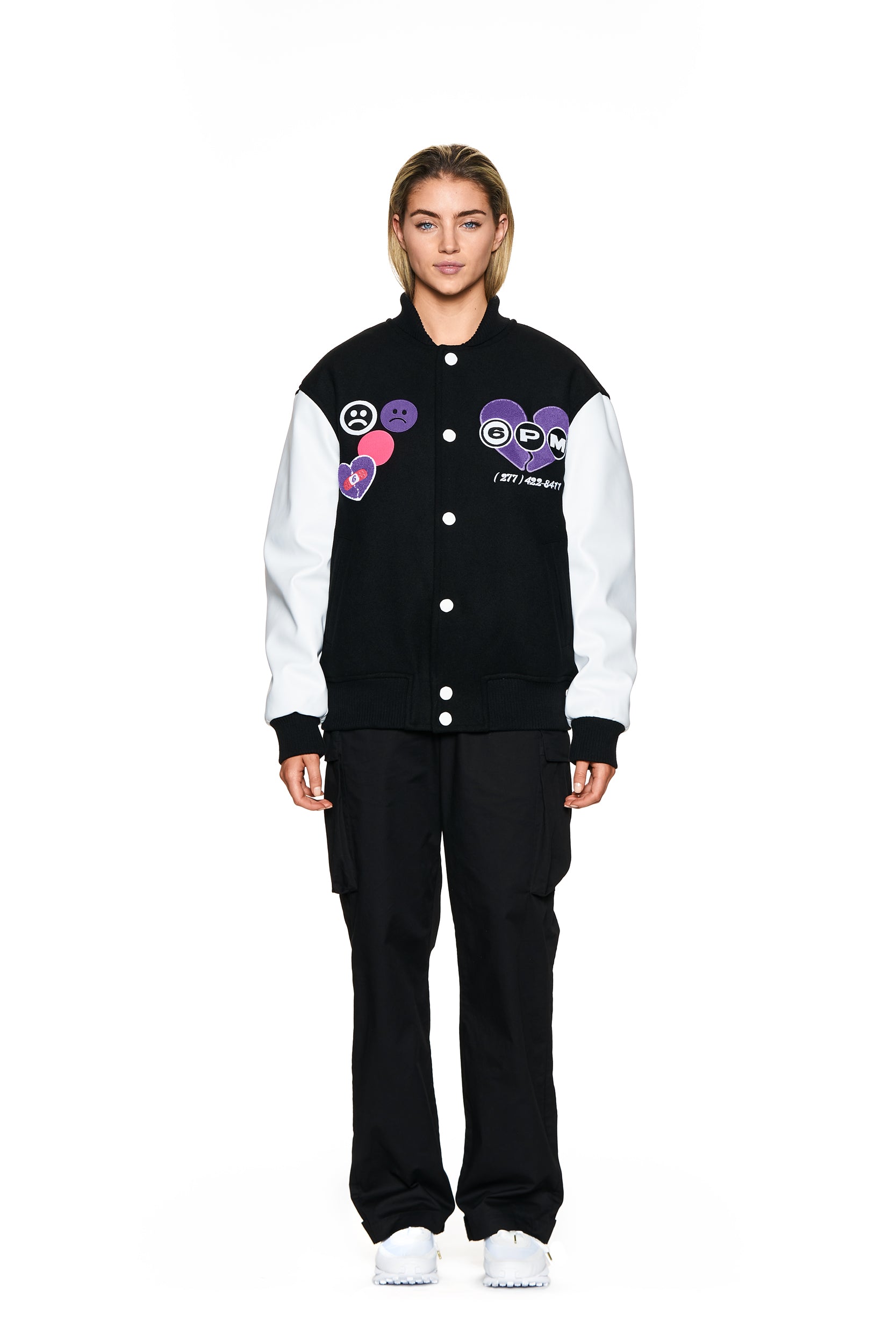 A person stands facing forward against a plain white background, wearing the 6PM COLLEGE JACKET BLACK with colorful patches, paired with black cargo pants and white sneakers.